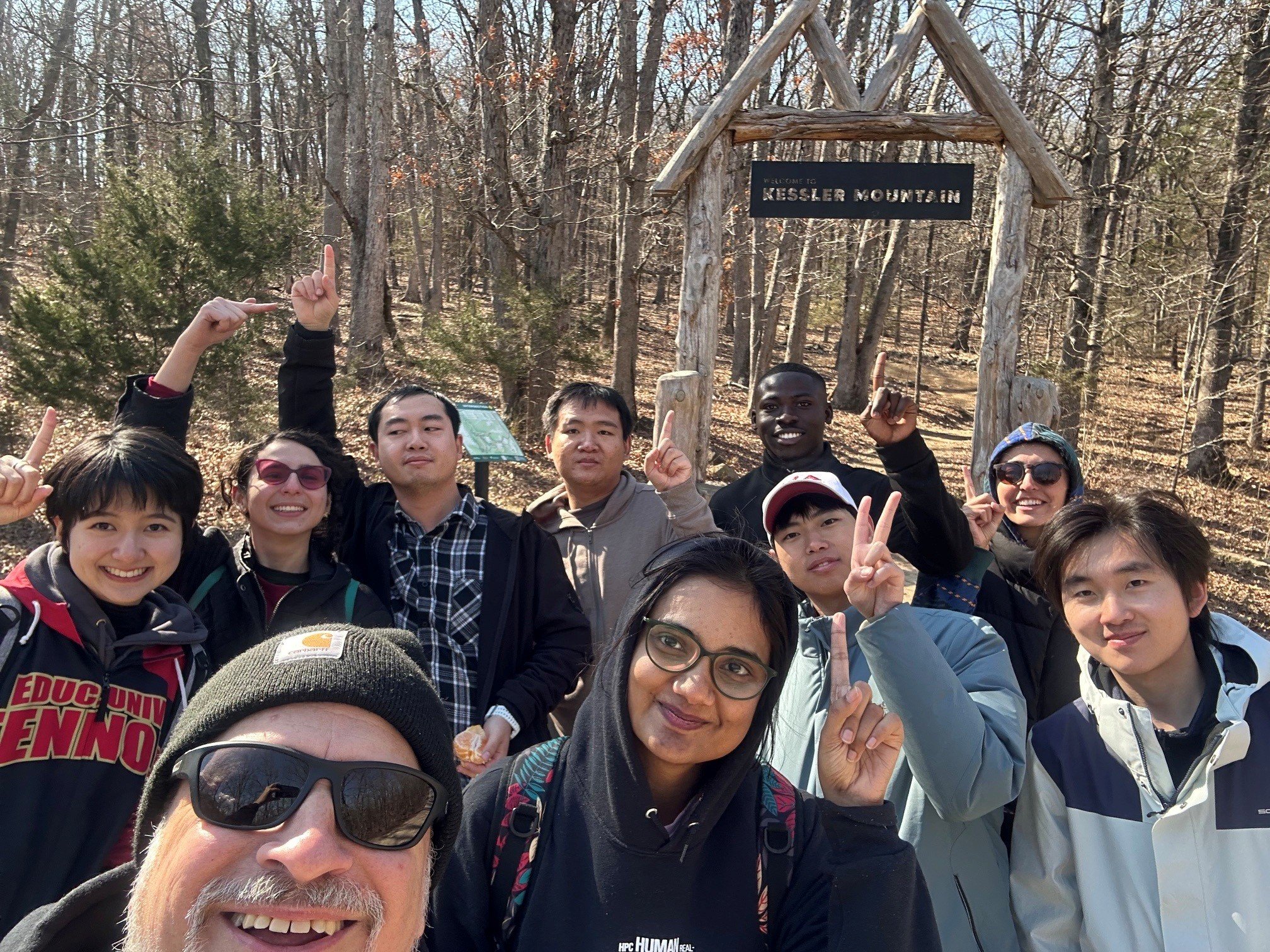 A group of diverse people (international students) and Joseph Poniatowski are taking a selfie in front of a wooden sign that reads 'Kessler Mountain' during a hike in a wooded area with leafless trees.