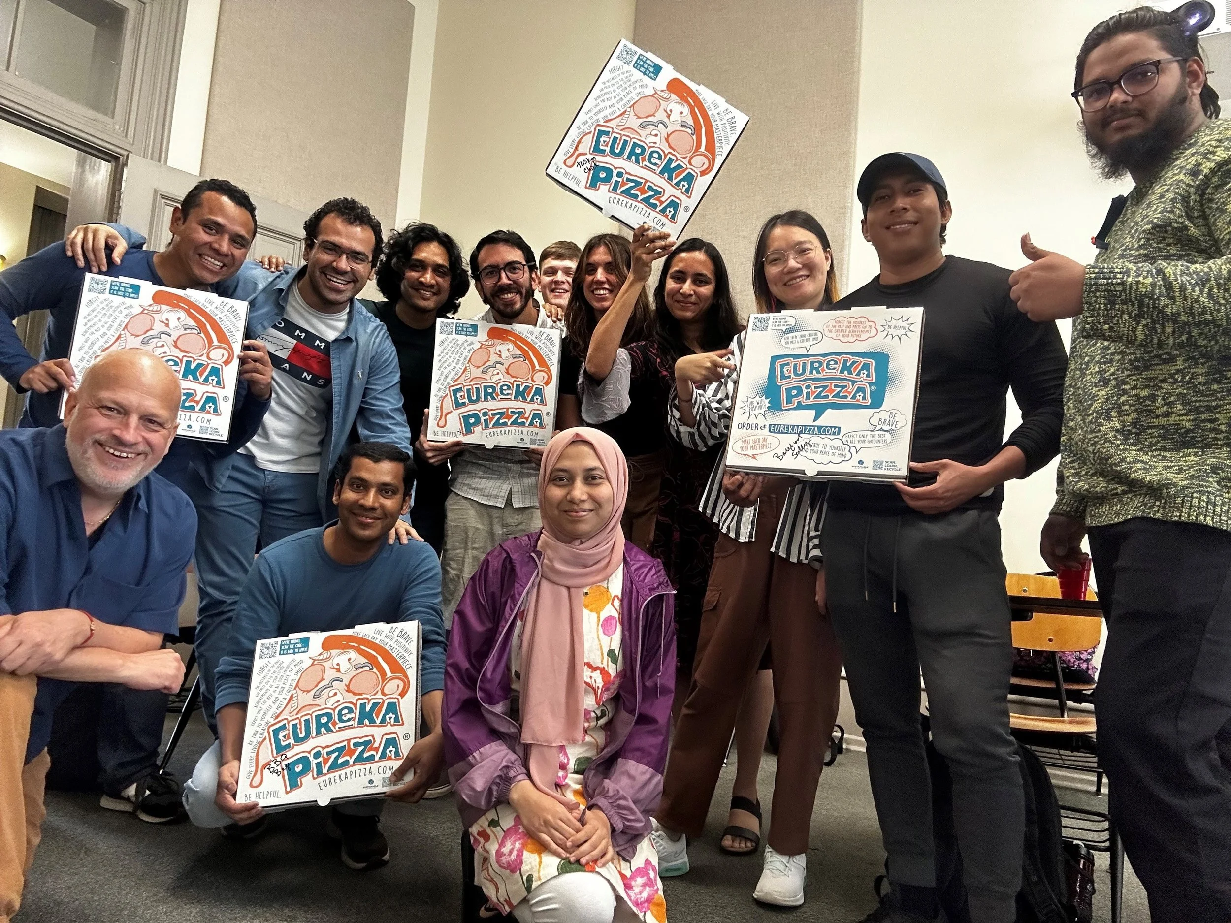 Group of diverse students and instructor Joseph Poniatowski posing indoors, holding signs with the Eureka Pizza logo, smiling, with some giving thumbs-up and others standing close together near the end of the semester at the University of Arkansas.