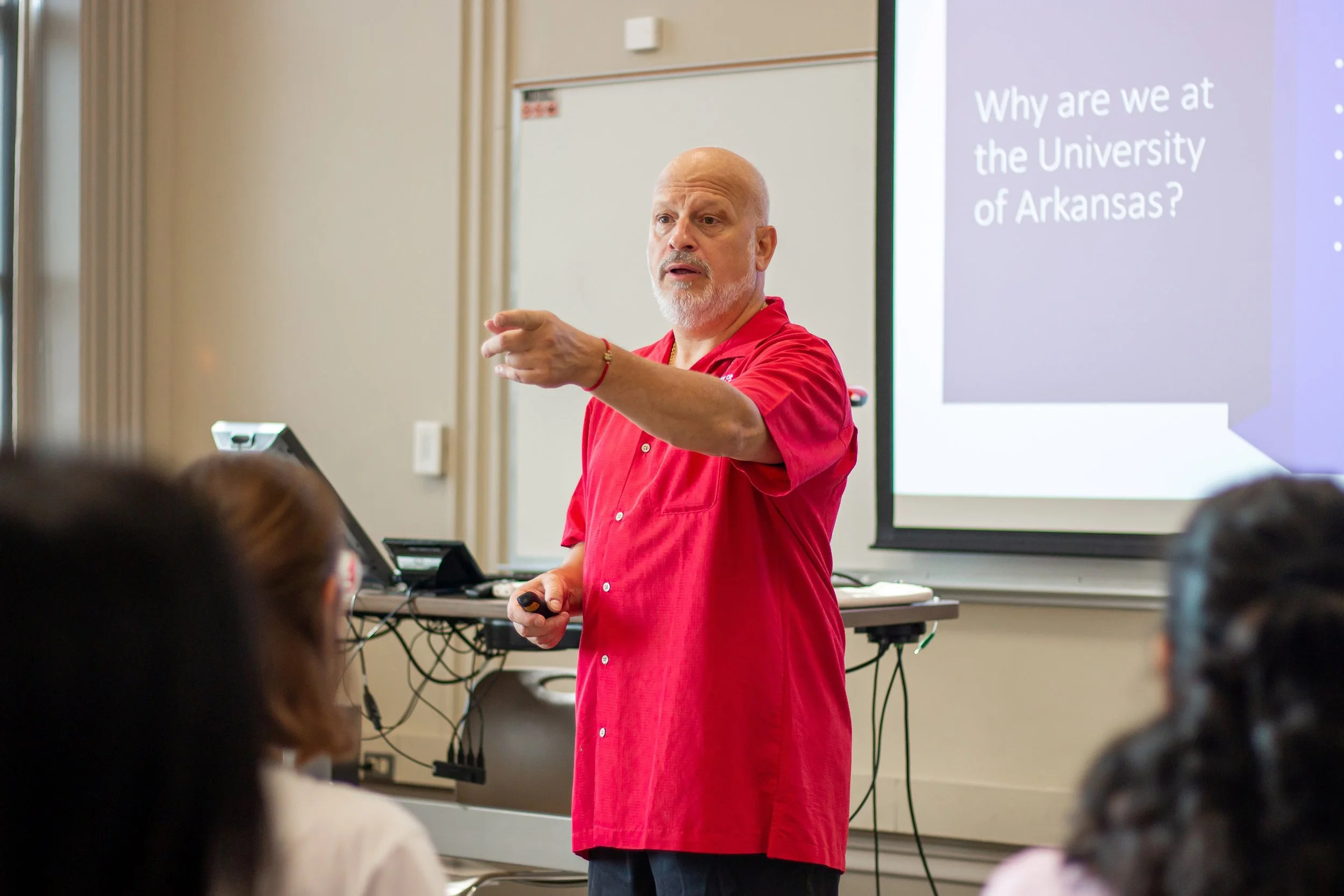 Joseph Poniatowski is wearing a red shirt and giving a presentation to a group of students in a classroom. There is a large screen behind him displaying text that reads 'Why are we at the University of Arkansas?'