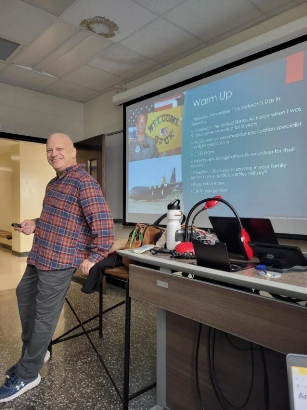 Joseph Poniatowski is standing and smiling in a classroom at the University of Arkansas near a table with electronics, with a presentation slide about Veteran's Day projected on the screen behind him.