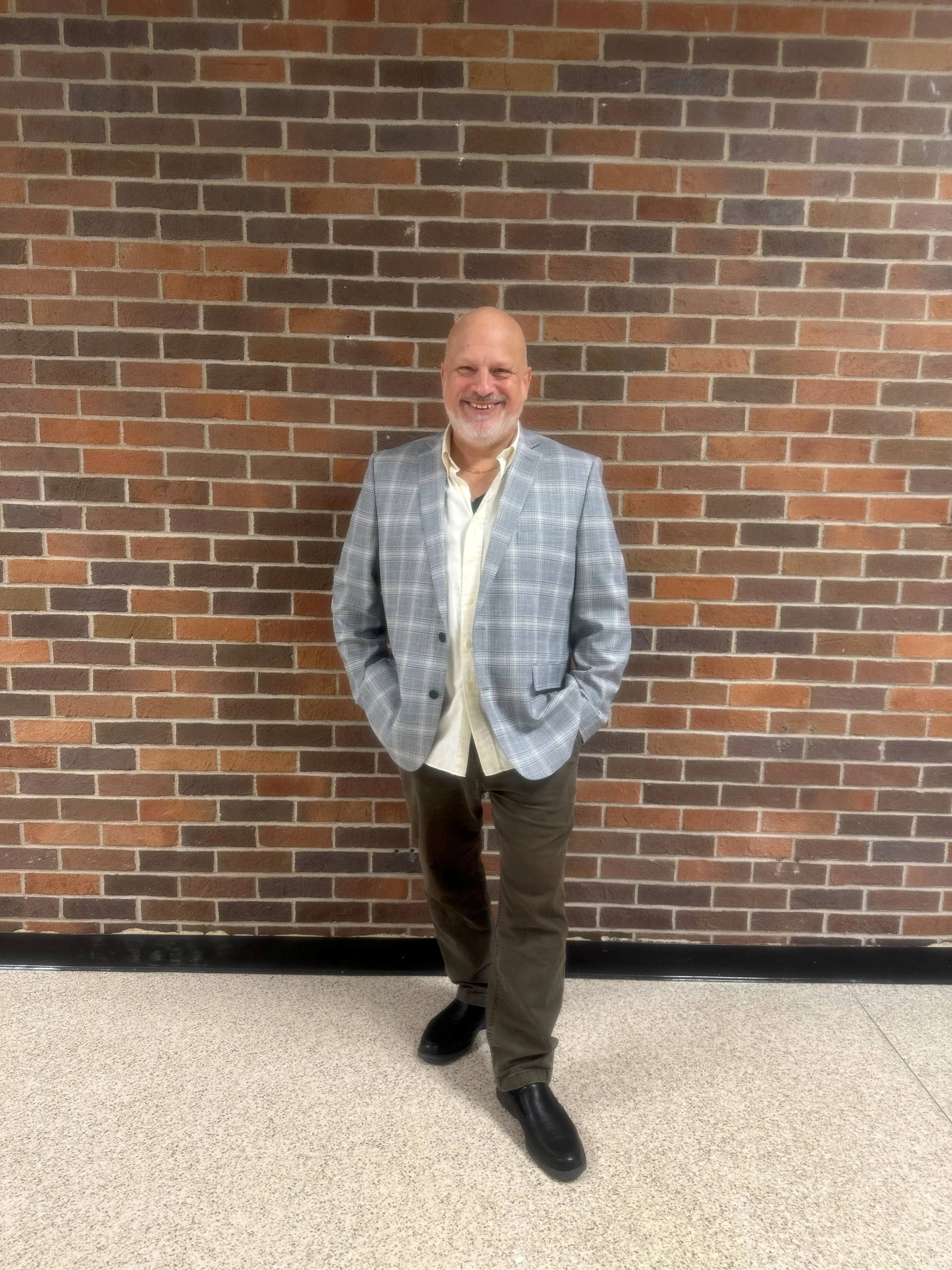 A man, Joseph Poniatowski, standing against a brick wall, wearing a light gray plaid blazer, light yellow shirt, dark pants, and black shoes, smiling with hands in pockets.