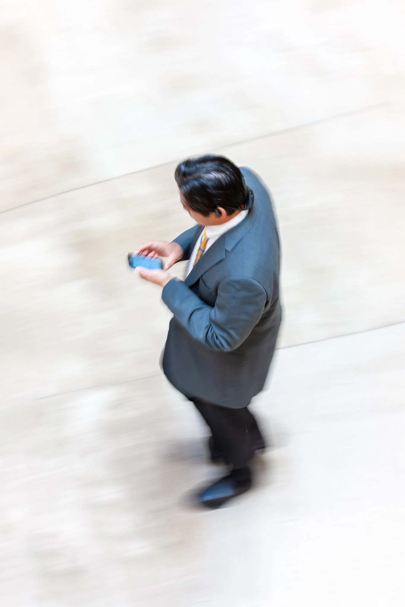 A high-angle photograph of a businessman in a suit standing on a stone floor, reading a message on his phone amid soft motion blur — from Ken Holden's Individual Personae collection.