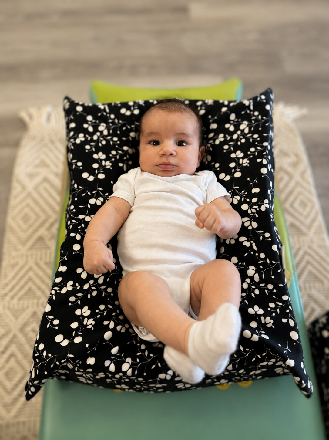 A baby lying on a black pillow with a white floral pattern, on a changing table with a patterned beige and white sheet, looking at the camera.