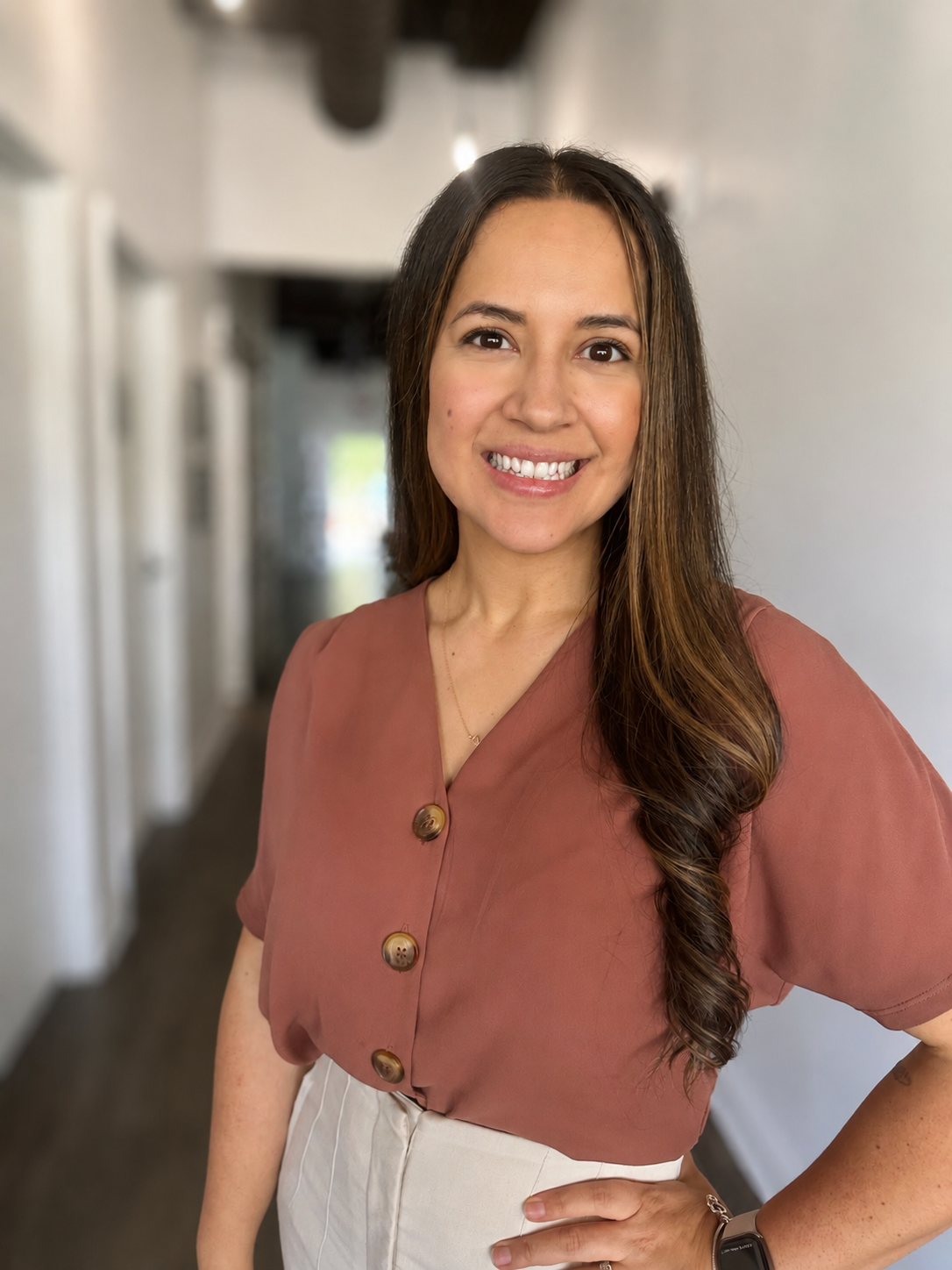 A woman with long brown hair wearing a rust-colored blouse with large buttons and a cream skirt standing in a hallway with white walls and wooden beams, smiling at the camera.