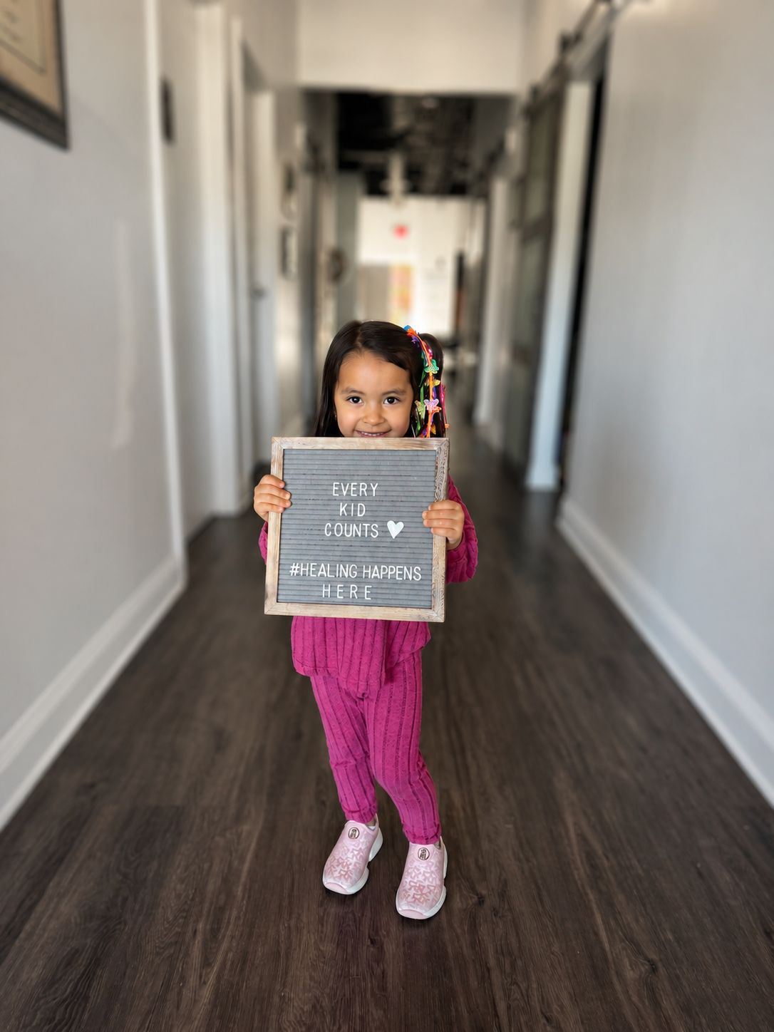 A young girl with colorful hair clips in her hair, wearing a pink outfit and white shoes, standing in a hallway and holding a sign that says 'Every kid counts. #healing happens here.'