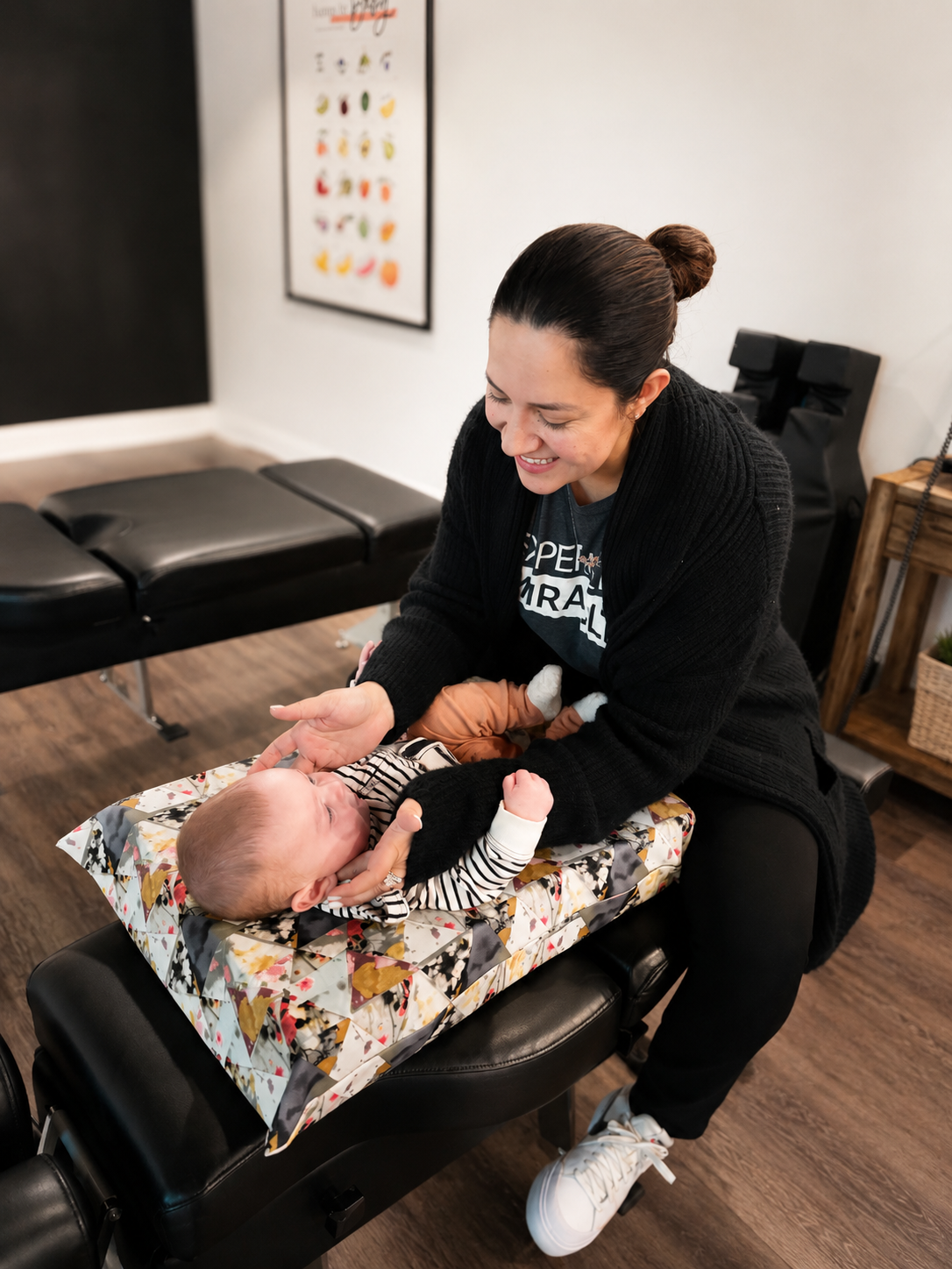 A smiling woman interacting with a baby lying on a padded table in a room with a massage table and a colorful poster on the wall.