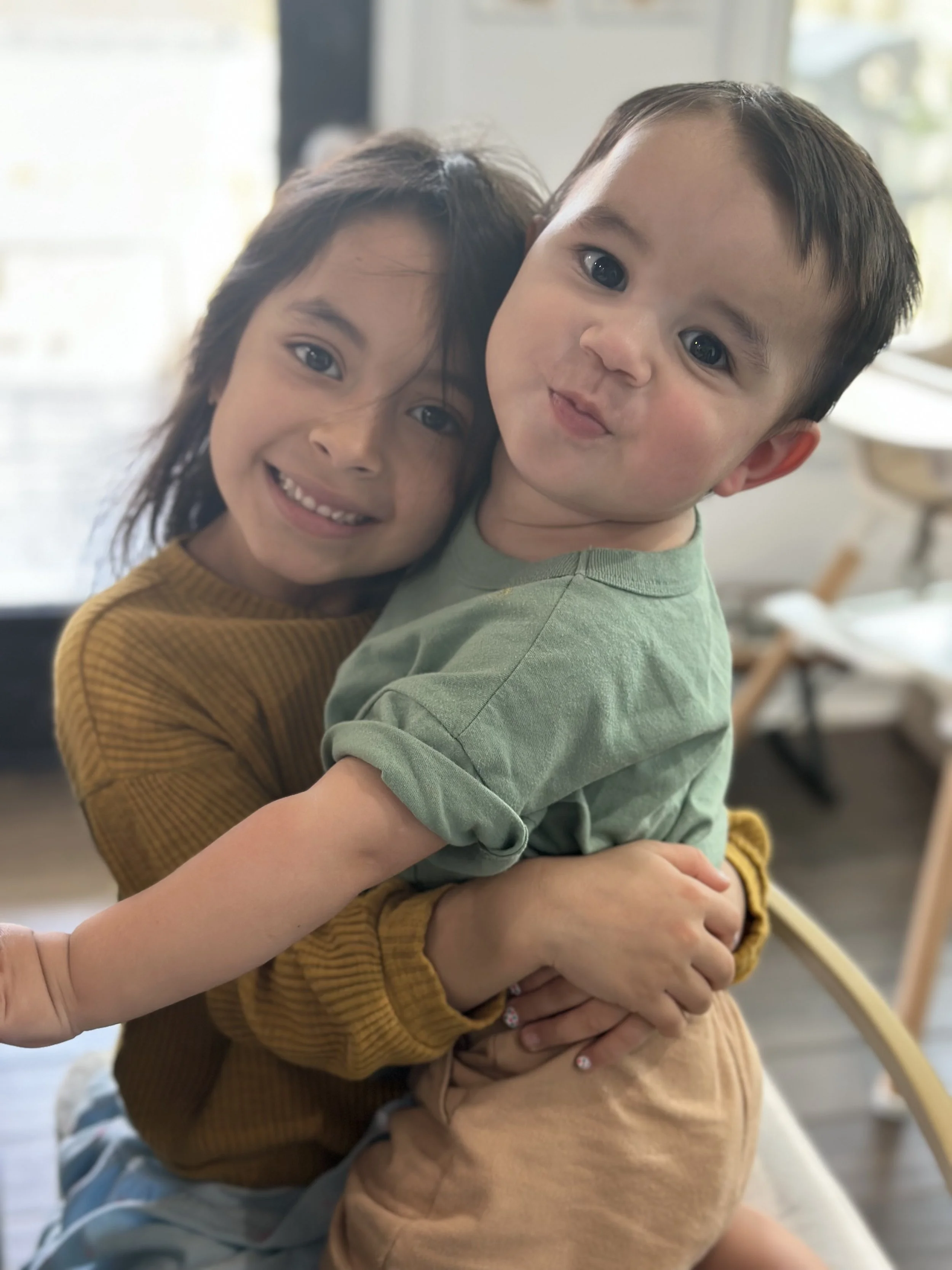 Two children, a girl and a boy, hugging each other indoors near a large window. The girl has long dark hair, is wearing a mustard-colored top, and is smiling. The boy has short dark hair, is wearing a green shirt, and has a playful expression sticking out his tongue.