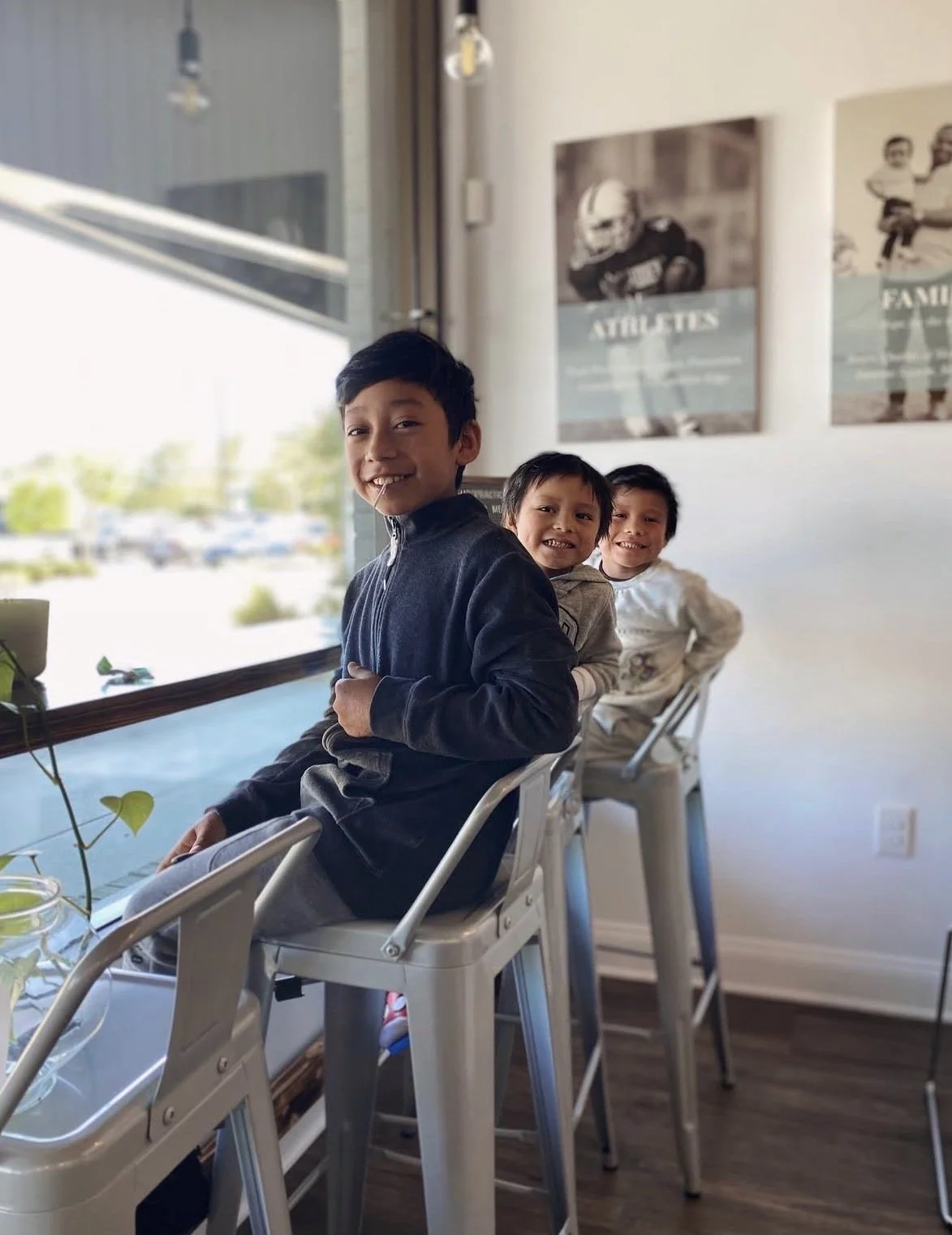 Three young boys sitting on chairs at a cafe or restaurant, smiling and looking at the camera. Behind them are large windows showing a parking lot and outdoors, with sports-themed posters on the wall.