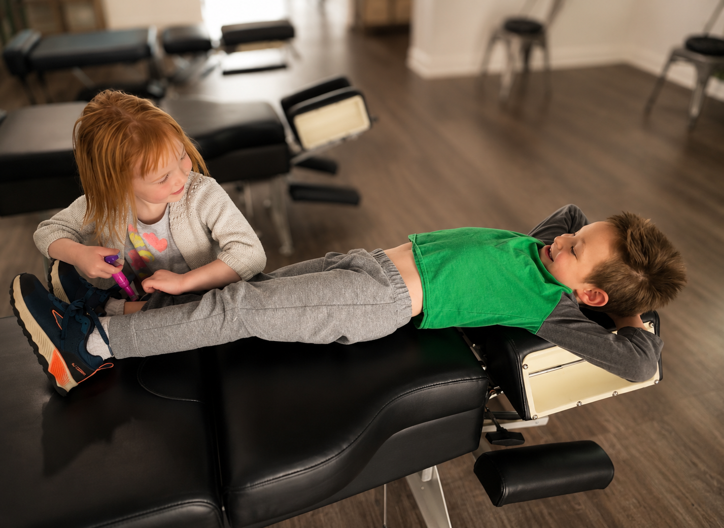 A young girl with red hair and a grey sweater is drawing on a boy with brown hair, lying on a medical examination table. The boy is wearing a green shirt and grey sweatpants, smiling and looking at the girl.