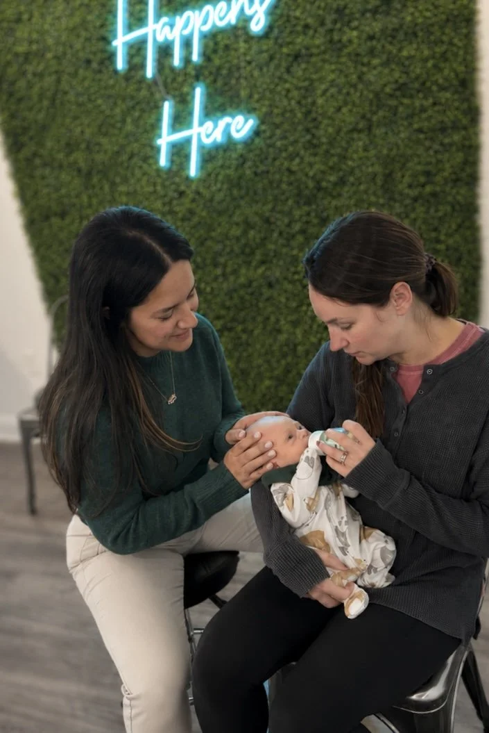 Two women holding a newborn baby in a cozy indoor setting with a green wall and a neon sign that says 'Happily Here'.