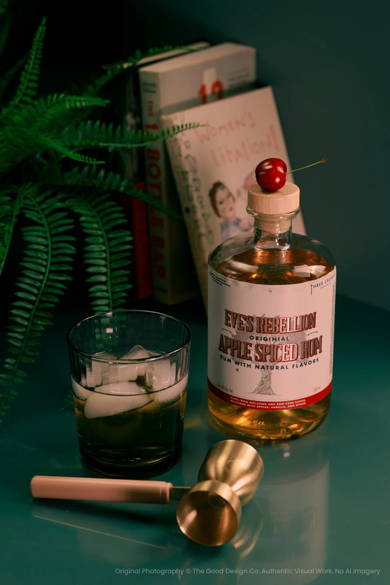 A bottle of Eve's Rebellion Original Apple Spiced Rum with a cherry on top of the bottle cap, a glass with ice and a drink, a pink straw, a gold ice scoop, and a green fern plant, with books and a note in the background.