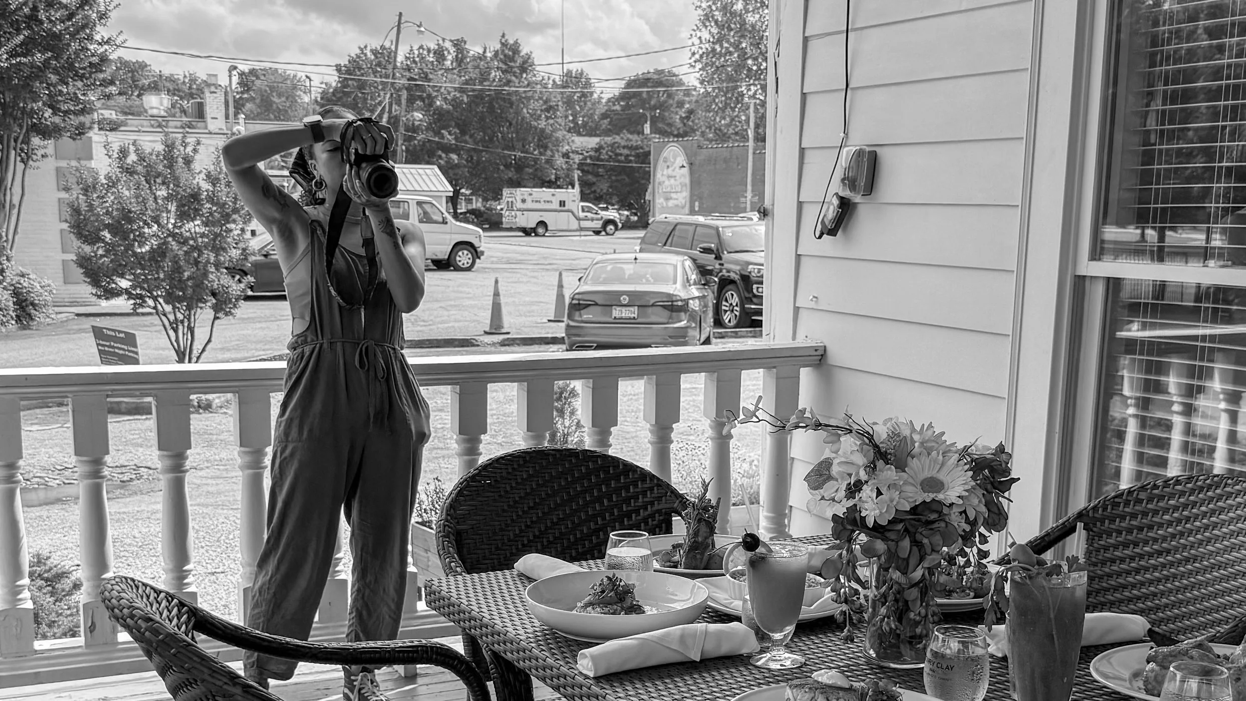 A woman standing on a porch taking a photo or looking through a camera. There are outdoor tables with flowers and glassware, and a view of a parking lot with cars and trees in the background.