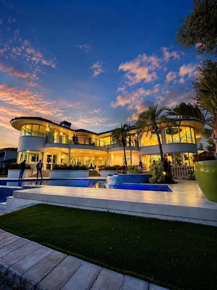 A luxurious modern house with large glass windows illuminated at sunset, surrounded by a swimming pool, palm trees, and a landscaped yard, with people socializing outside.