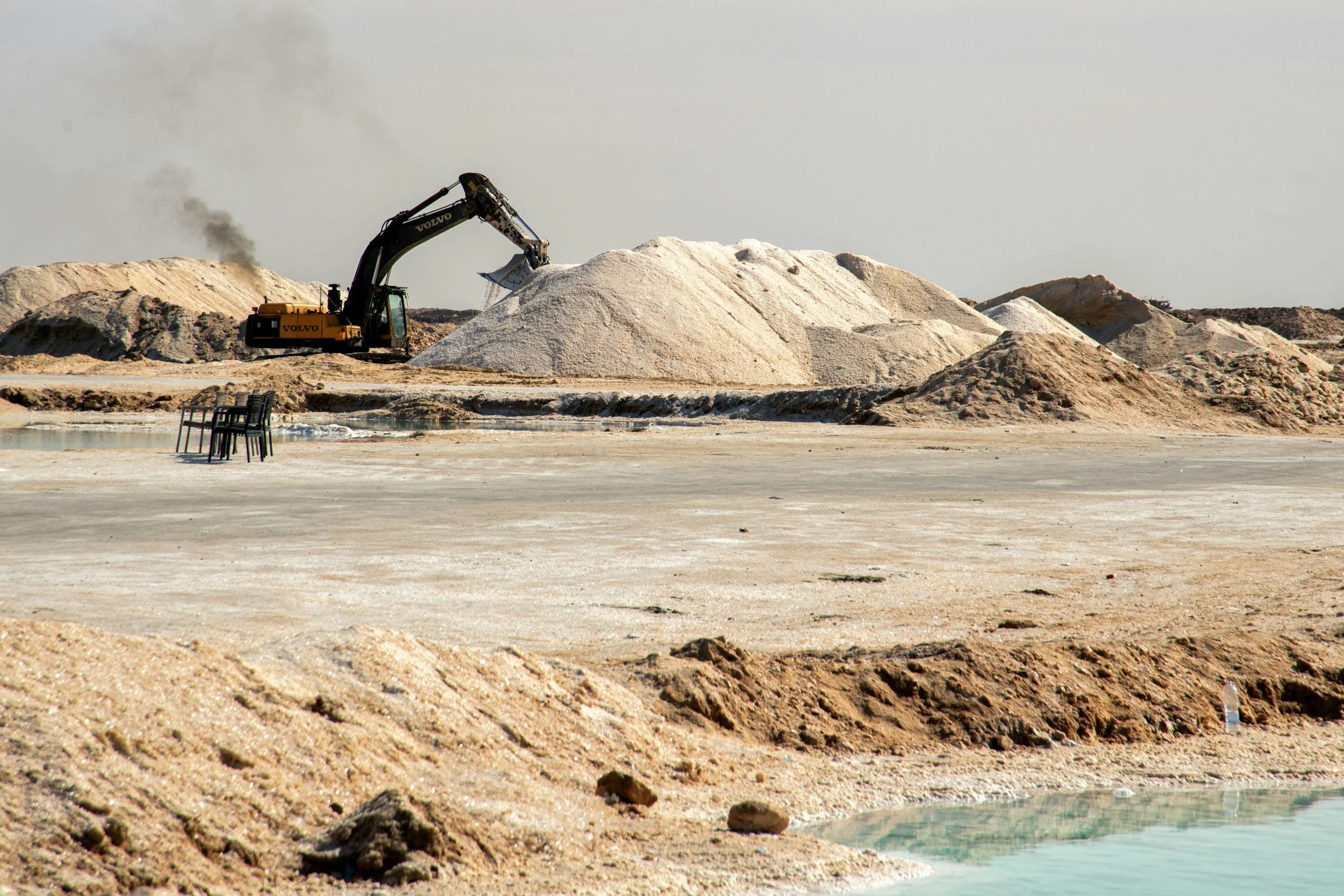 Une excavatrice jaune creuse une montagne de sable dans un paysage désertique, avec un peu de vapeur ou de fumée s'échappant au loin, et quelques chaises noires au premier plan.