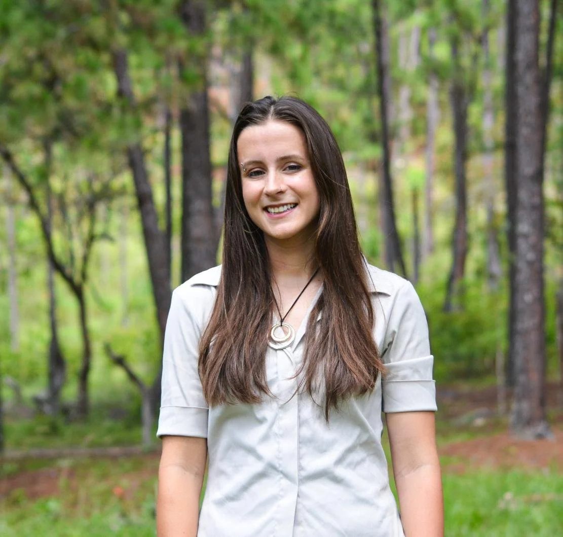 A woman smiling outdoors in front of a large tree with green leaves, wearing a teal blazer over a black top.
