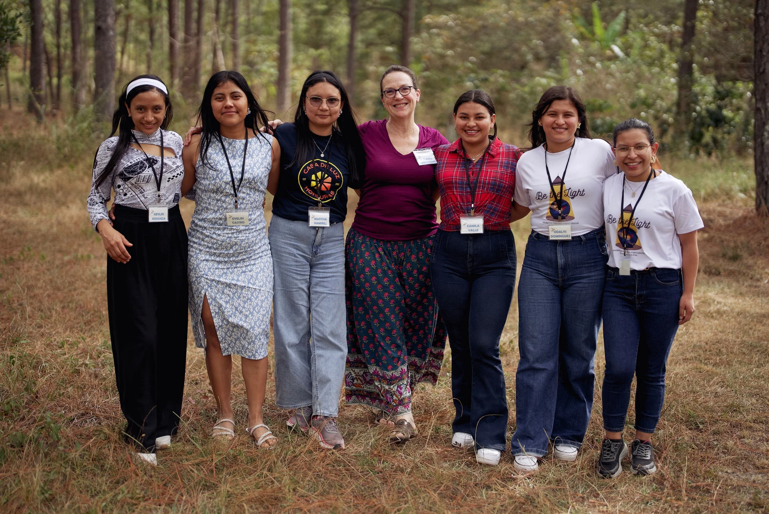 Group of seven women standing together outdoors in a forest, smiling for a photo.