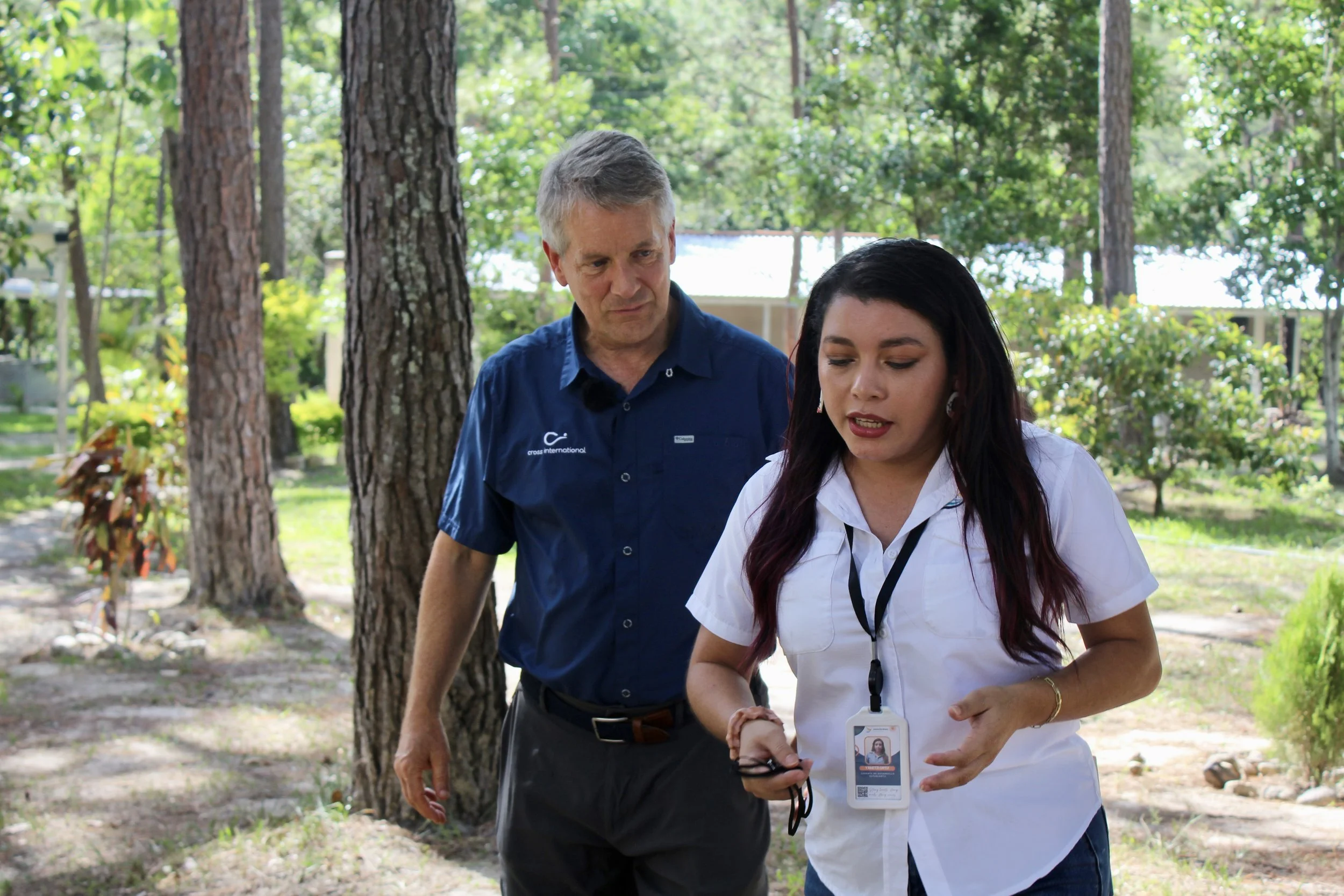 A man and a woman walk outdoors in a wooded area. The man is wearing a dark blue shirt, and the woman, dressed in a white shirt, has identification and a lanyard around her neck.
