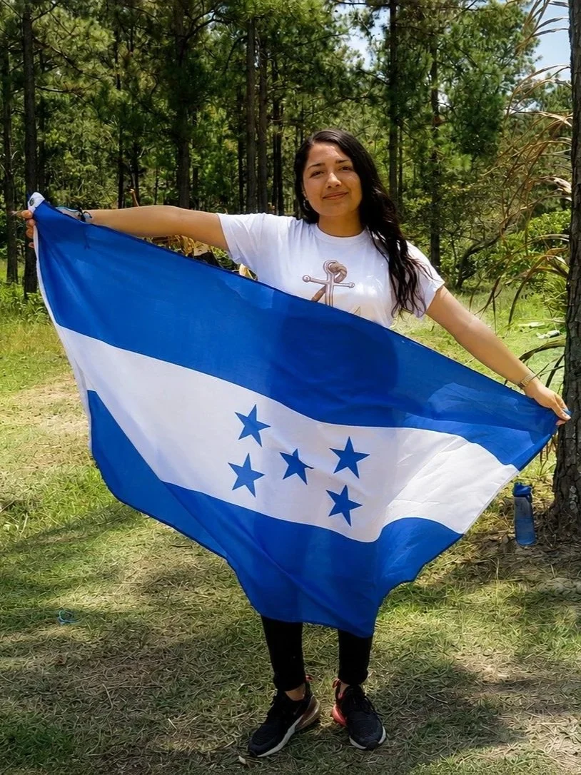 A woman standing outdoors in a wooded area, holding a blue and white Honduras flag with five blue stars arranged in a semi-circle, wearing a white T-shirt with an anchor design, black pants, and black sneakers.