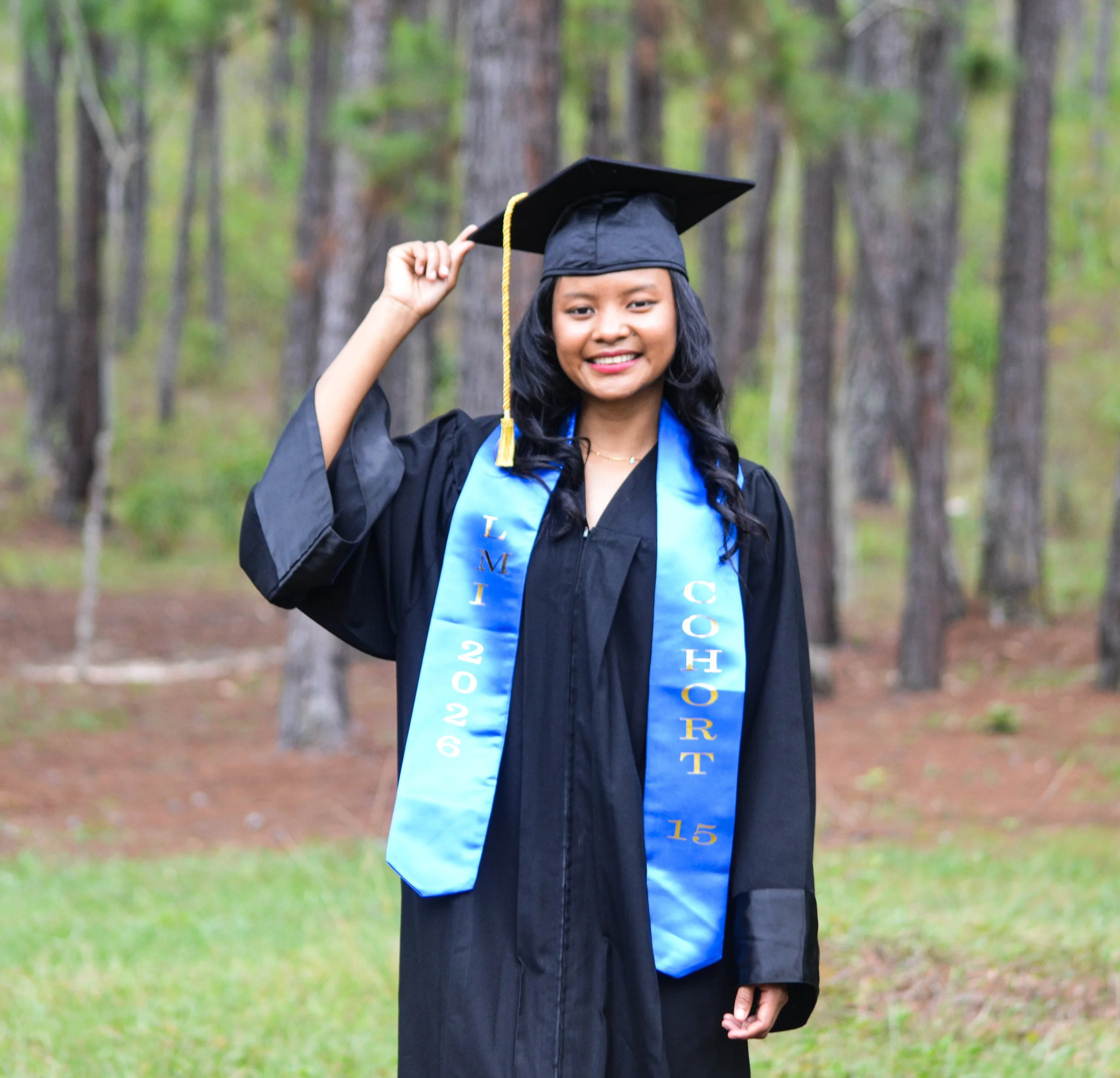 A young woman in a black graduation gown and cap with a gold tassel, standing outdoors in a wooded area, smiling and adjusting her cap. She is wearing a blue stole with gold lettering, indicating a graduation ceremony.