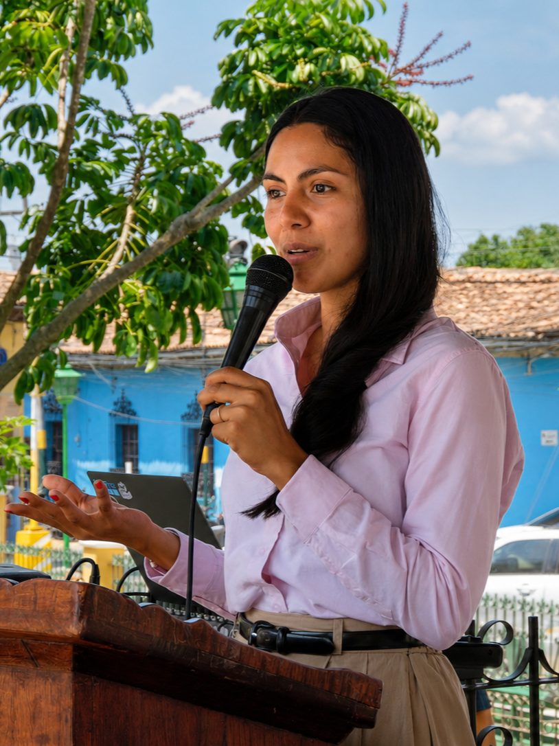 A woman with long dark hair, wearing a light pink blouse, speaking into a microphone outdoors next to a wooden podium, with trees and a blue building in the background.