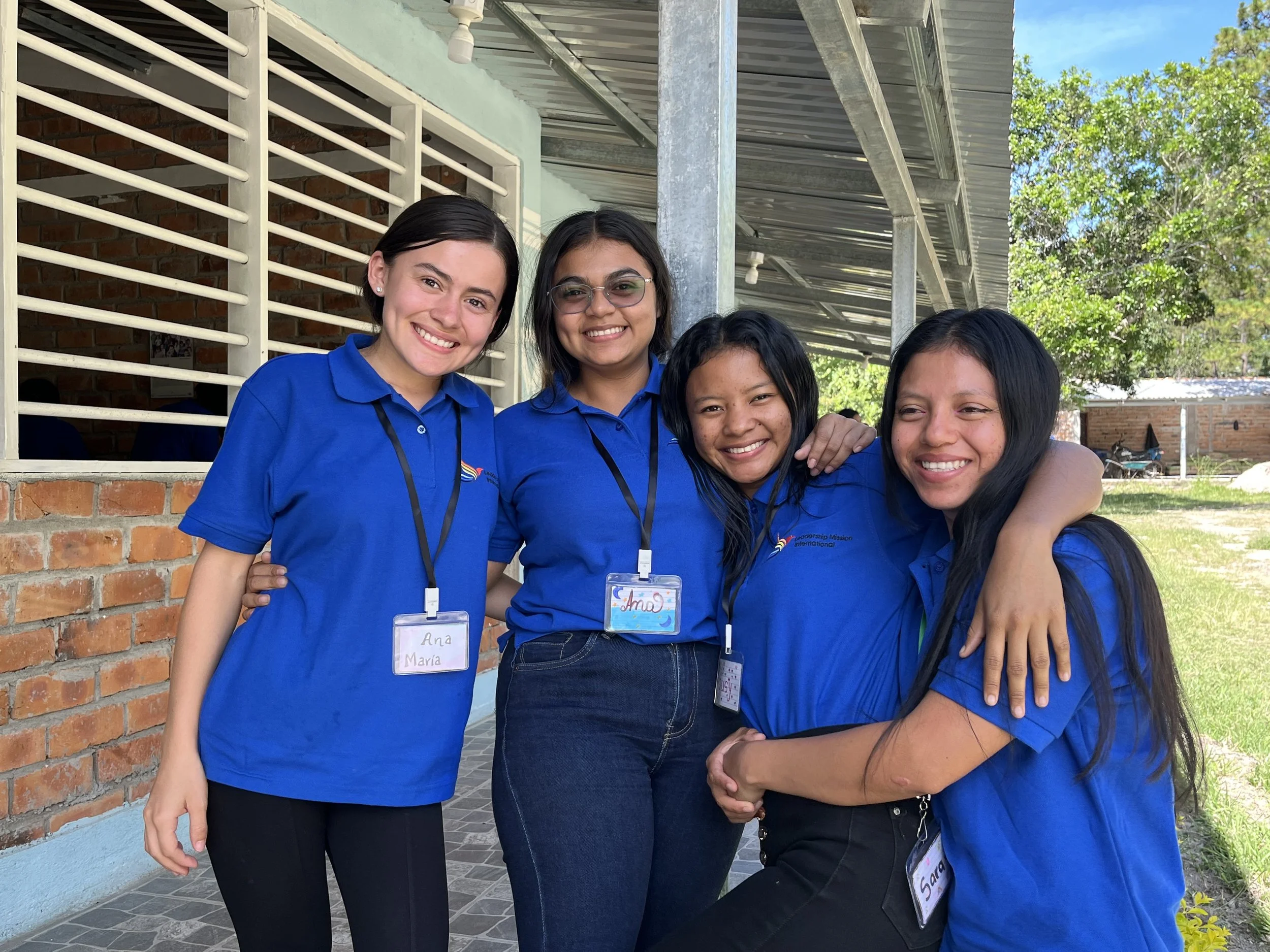 Four young women wearing blue shirts standing close together outside near a brick and painted building, smiling and embracing each other. They are wearing identification badges around their necks.
