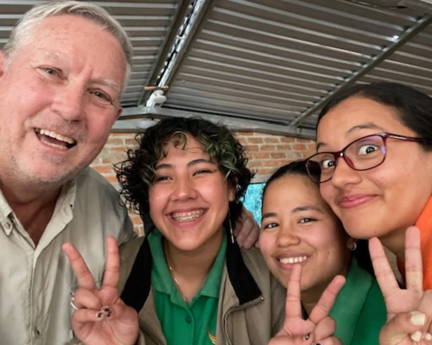 Group of four smiling people taking a selfie, making peace signs with their hands, indoors with a brick wall and metal roof in the background.