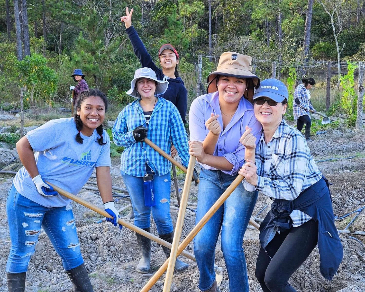 Group of women and girls working outdoors planting trees or plants in a forested area, holding gardening tools, and smiling at the camera.