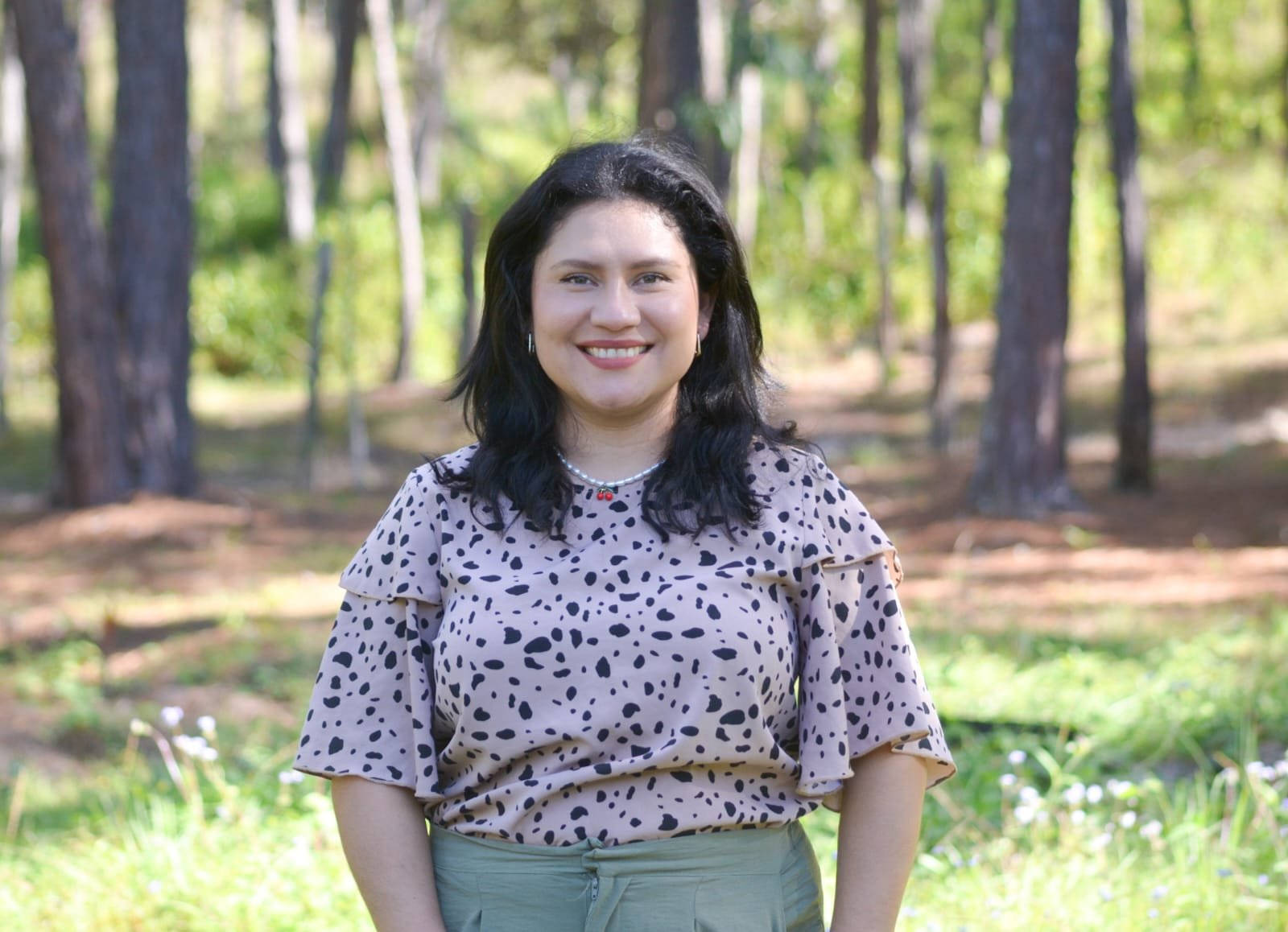A woman with dark hair smiling outdoors in a forest setting, wearing a beige dress with black spots, a pearl necklace, and earrings.