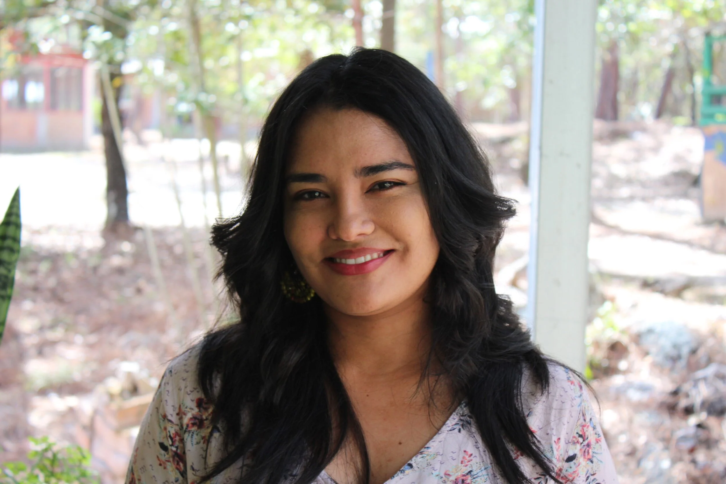 A woman with long black hair smiling outdoors during daytime with a background of trees and nature.