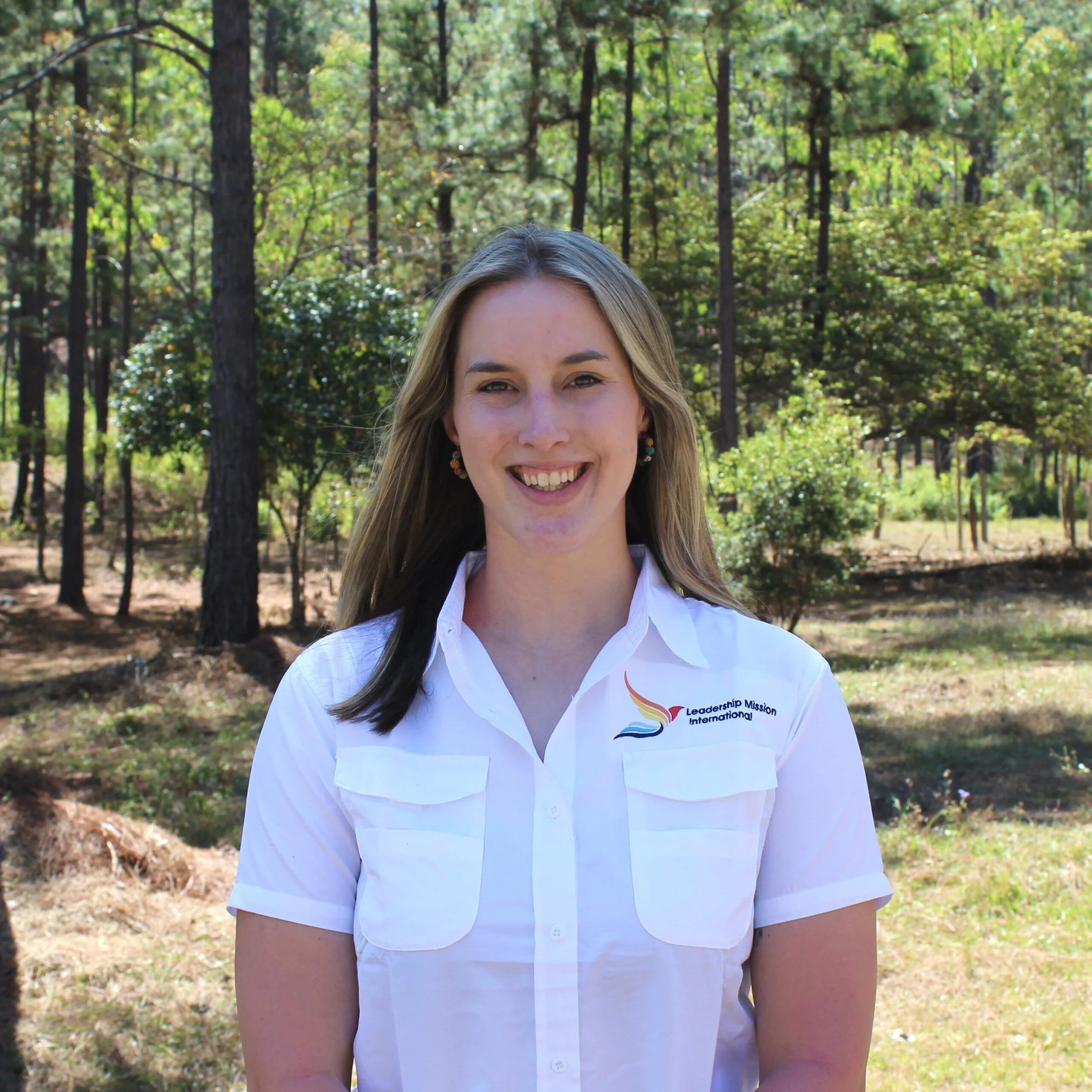 A smiling woman with long wavy blonde hair wearing a white shirt with a logo that reads 'Leadership Mission International' standing outdoors among trees and greenery.