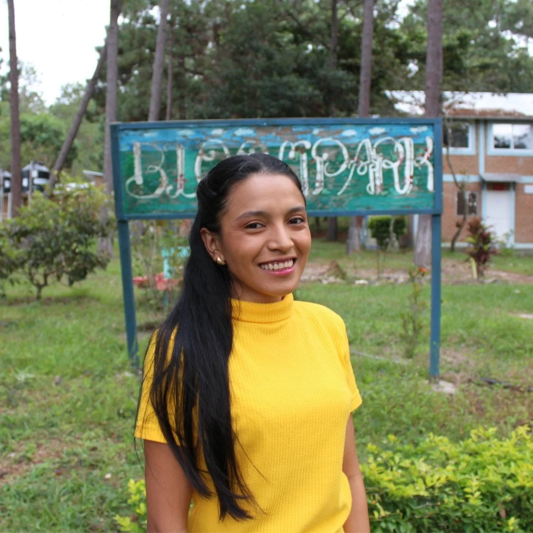 A woman with long black hair wearing a yellow turtleneck sweater, standing outdoors in front of a green, weathered sign with faded writing, with trees and a building in the background.