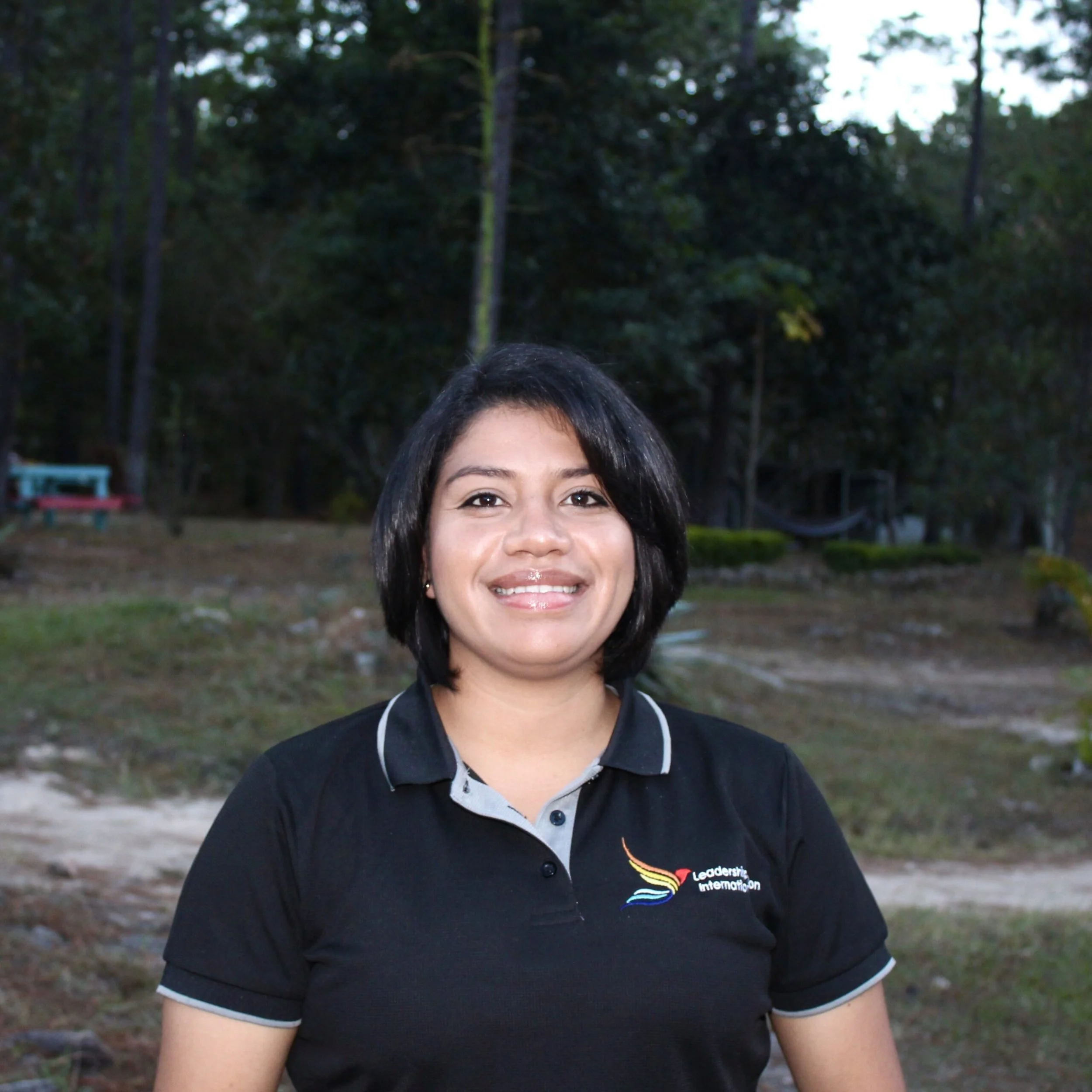 Close-up of a smiling woman with black hair wearing a black polo shirt with a colorful logo, standing outdoors in a wooded area during daytime.