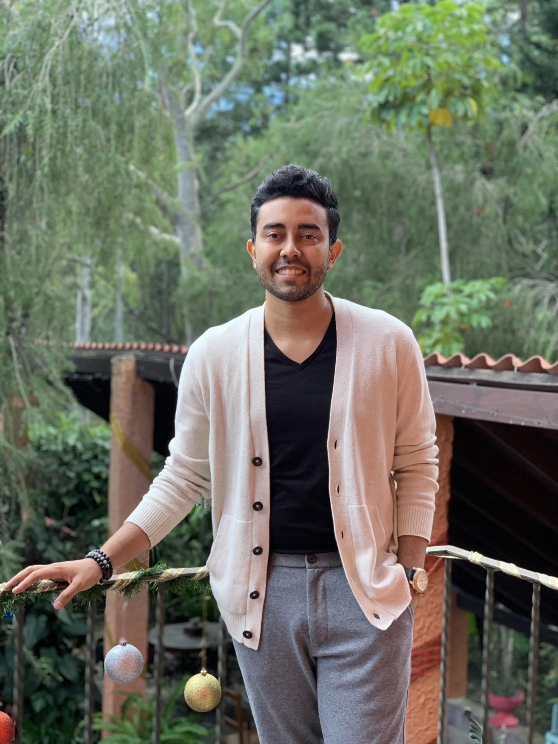 A young man with short dark hair and a beard, smiling, standing outdoors in front of green trees. He is wearing a light beige cardigan over a black shirt, gray pants, a watch on his left wrist, and beaded bracelets on his right wrist. He is holding onto a holiday garland decorated with colorful Christmas ornaments, including a glittery silver and gold ball.