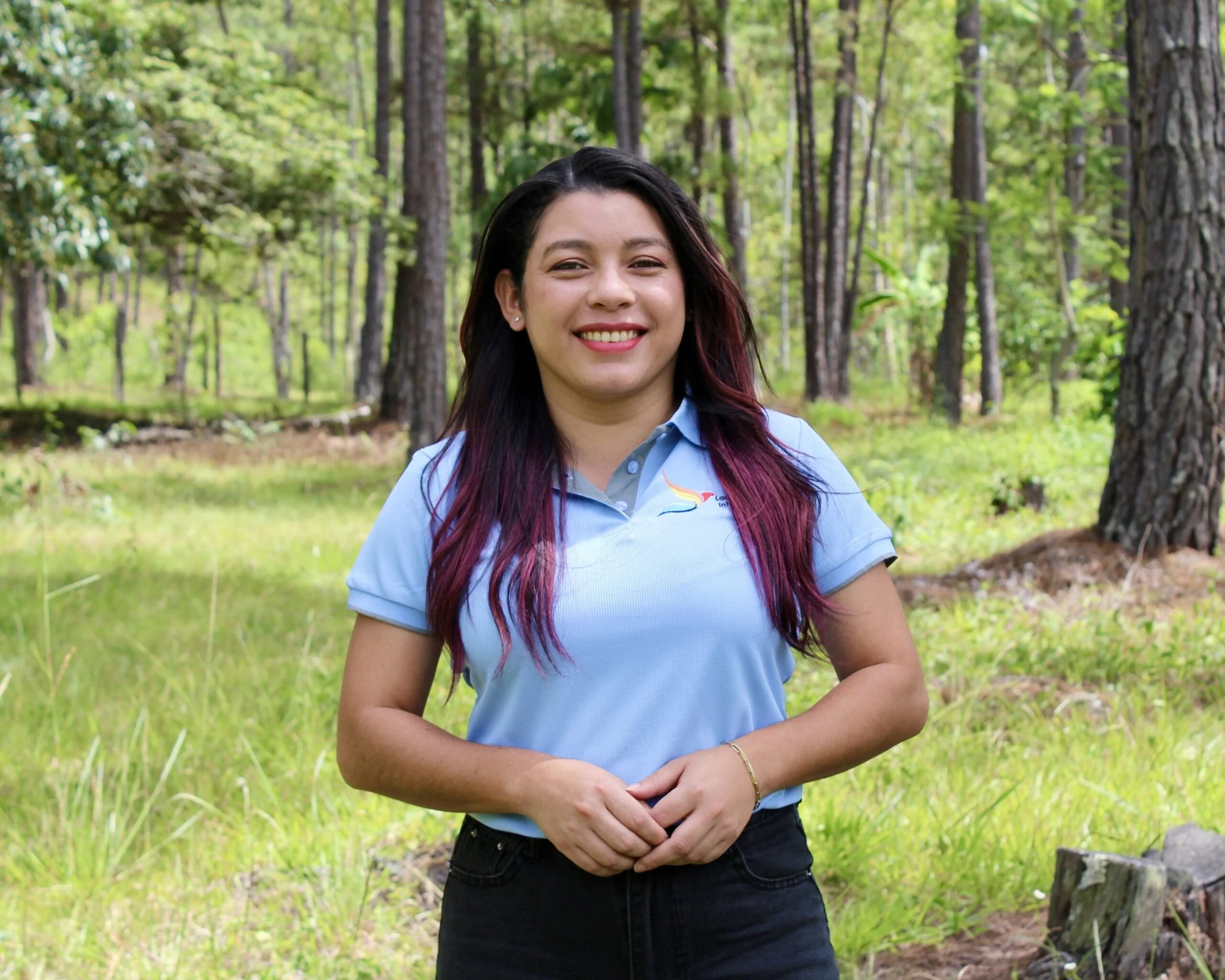 A smiling woman with long dark hair with purple highlights, wearing a light blue polo shirt, standing outdoors in a green forested area with trees and grass.