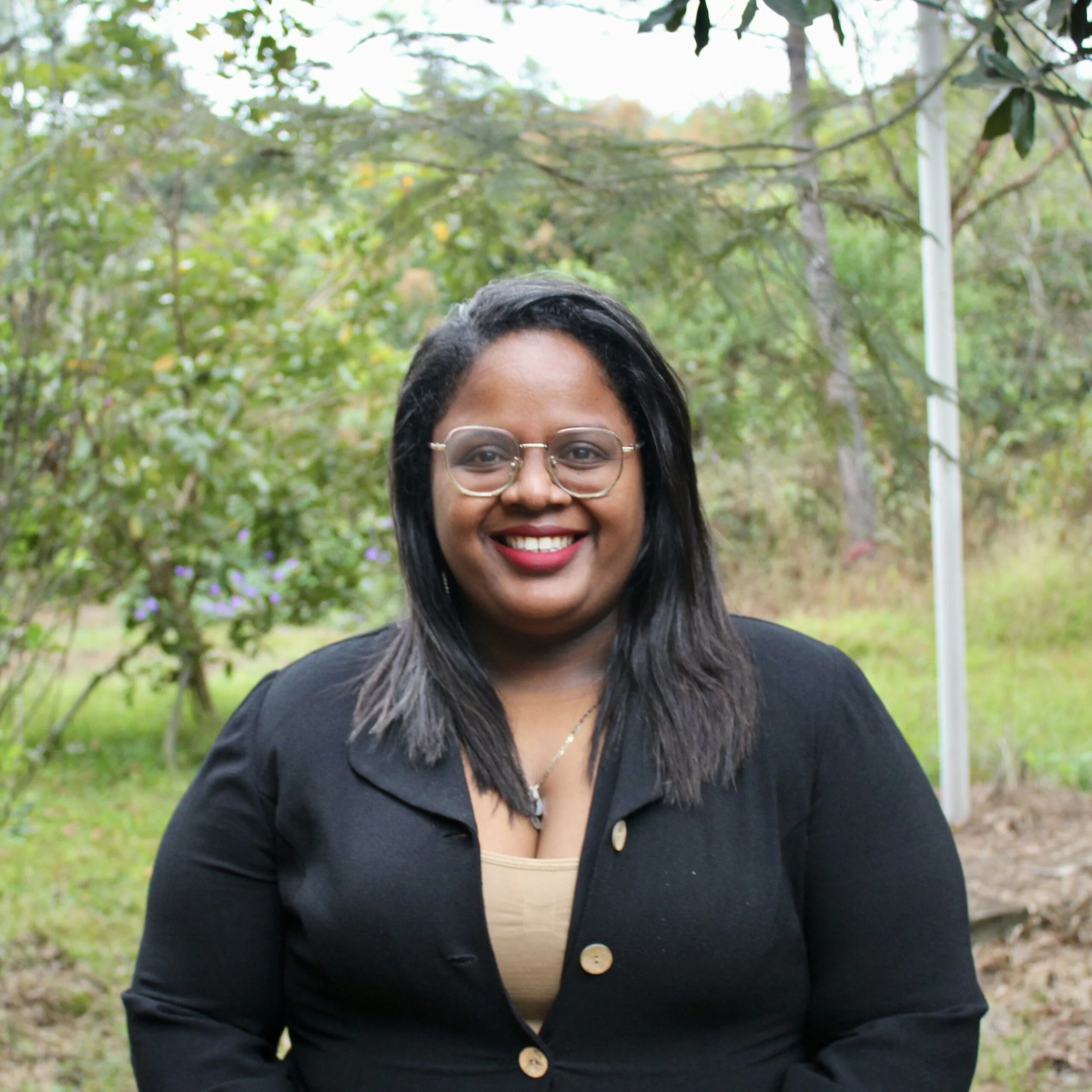 Close-up of a smiling woman with glasses and long black hair, wearing a black jacket and beige top, standing outdoors in a park with green trees and foliage.
