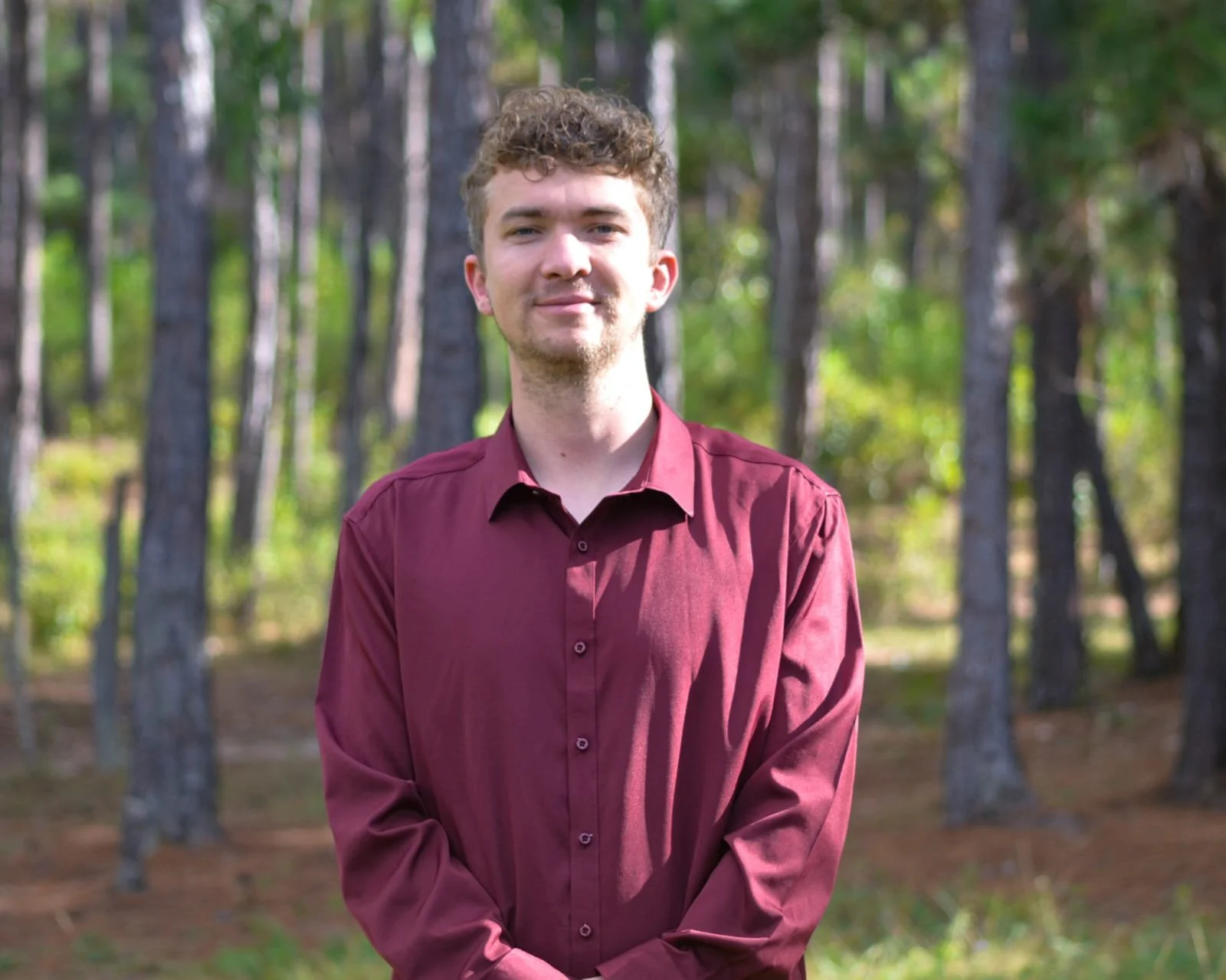 A young man with curly hair wearing a maroon shirt standing outdoors in a wooded area.