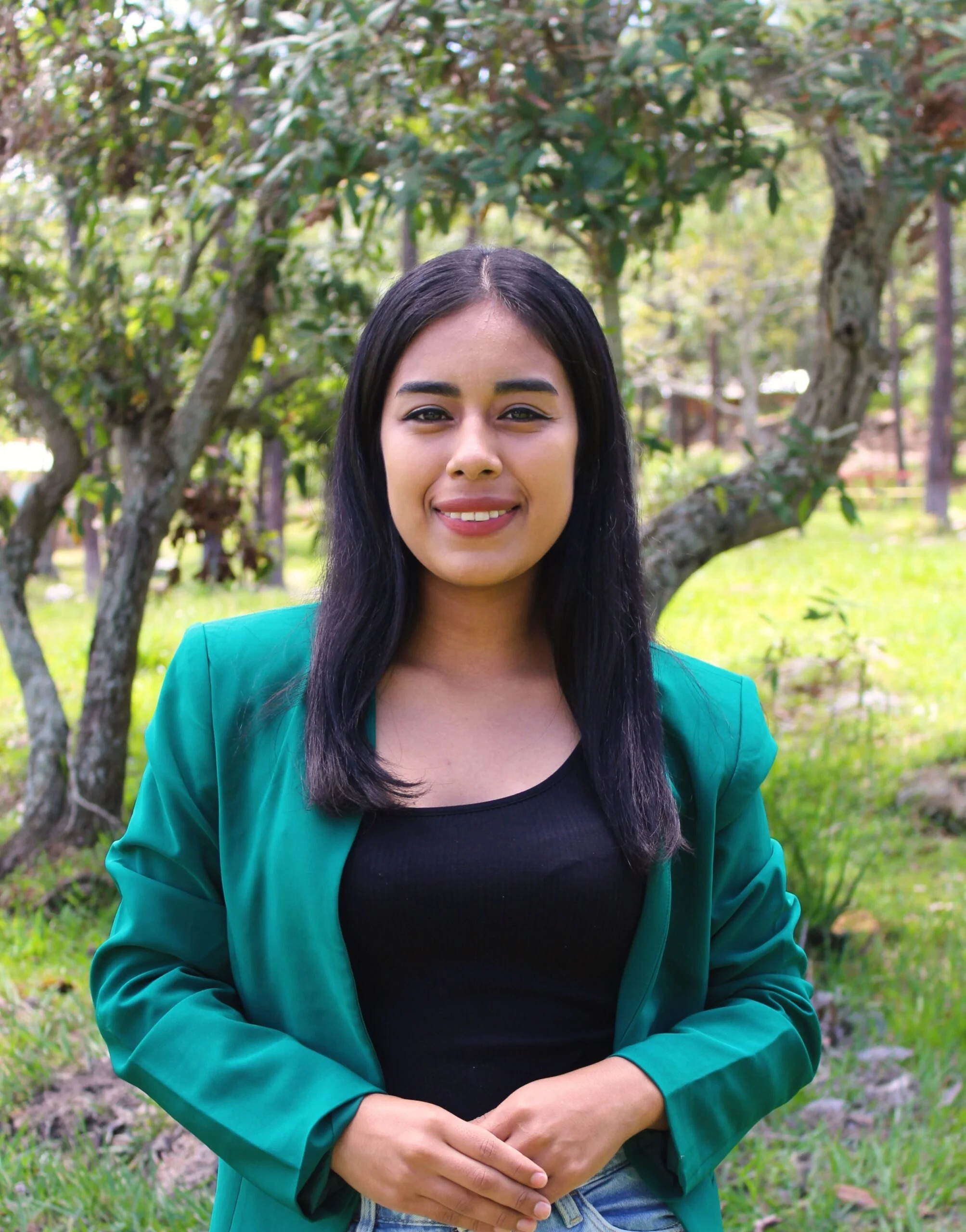 A woman smiling outdoors in front of a large tree with green leaves, wearing a teal blazer over a black top.