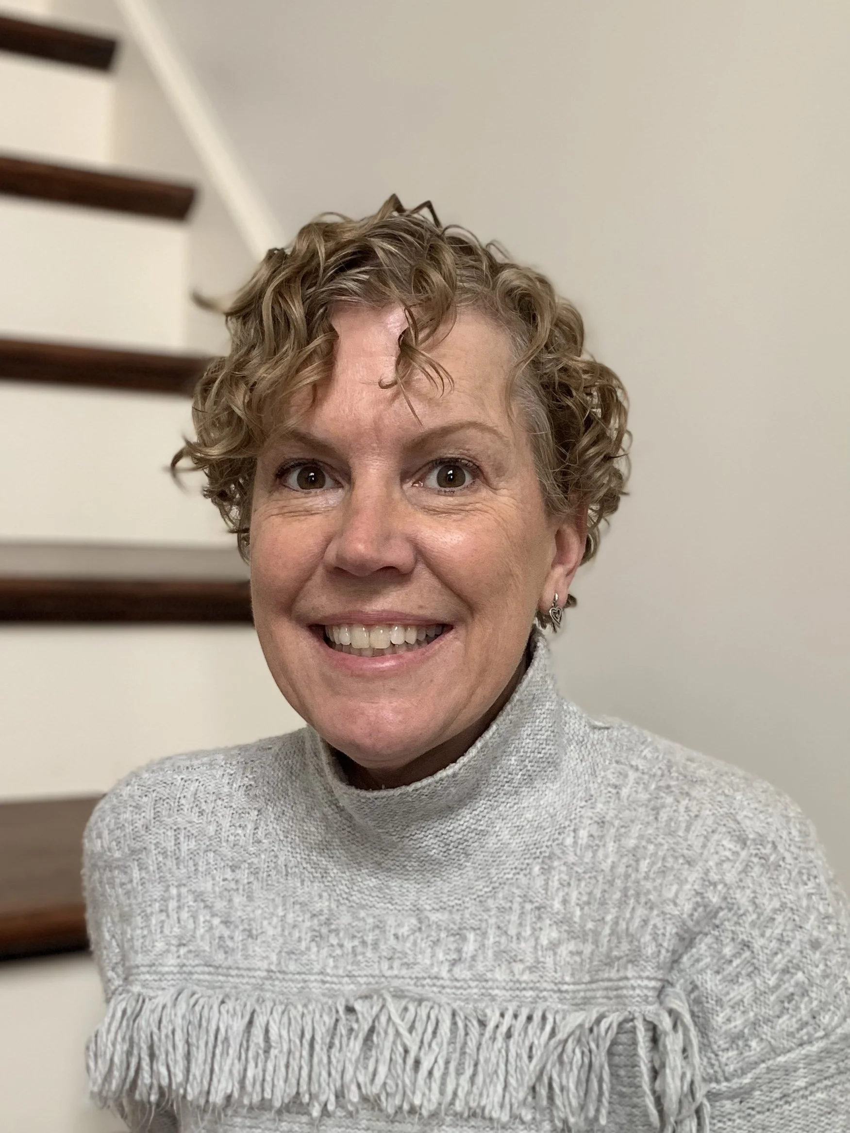 A smiling middle-aged woman with curly short hair, wearing a light gray sweater with fringes, standing indoors against a beige wall with wooden stairs in the background.