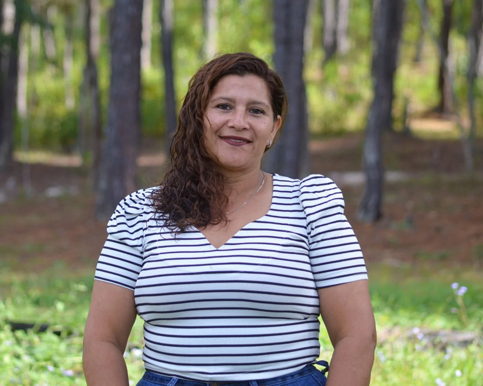 A woman with curly brown hair, wearing a striped white and navy shirt, standing outdoors in a wooded area with green trees and grass in the background.