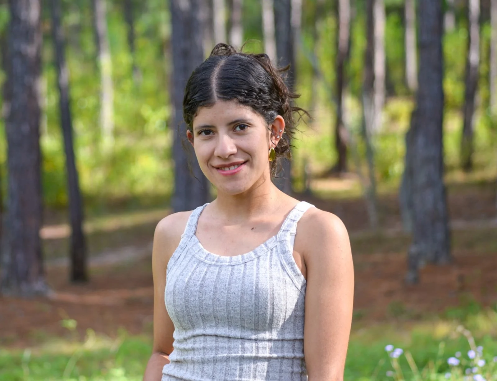 A young woman with dark, wavy hair and multiple earrings, wearing a light gray sleeveless top, standing outdoors in a wooded area with trees and green foliage in the background.