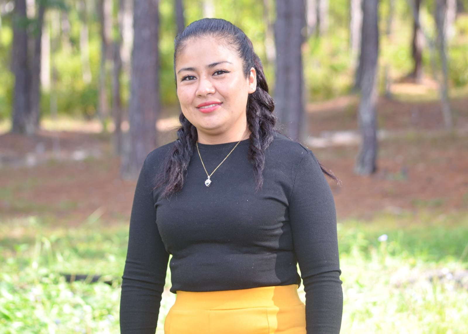 A woman with dark hair tied in two braids, wearing a black long-sleeve shirt and a yellow skirt, standing outdoors in a wooded area with trees and green foliage in the background.