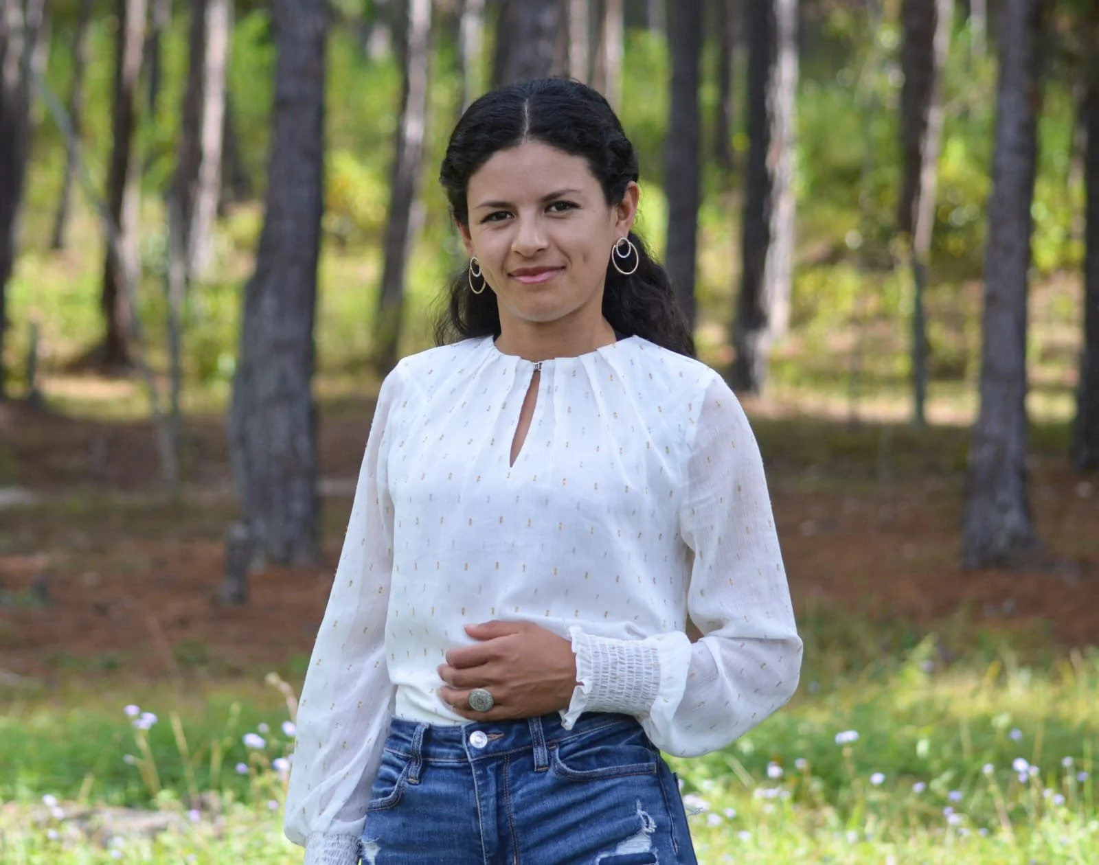 A woman with dark hair wearing a white blouse and jeans standing in a wooded outdoor area.