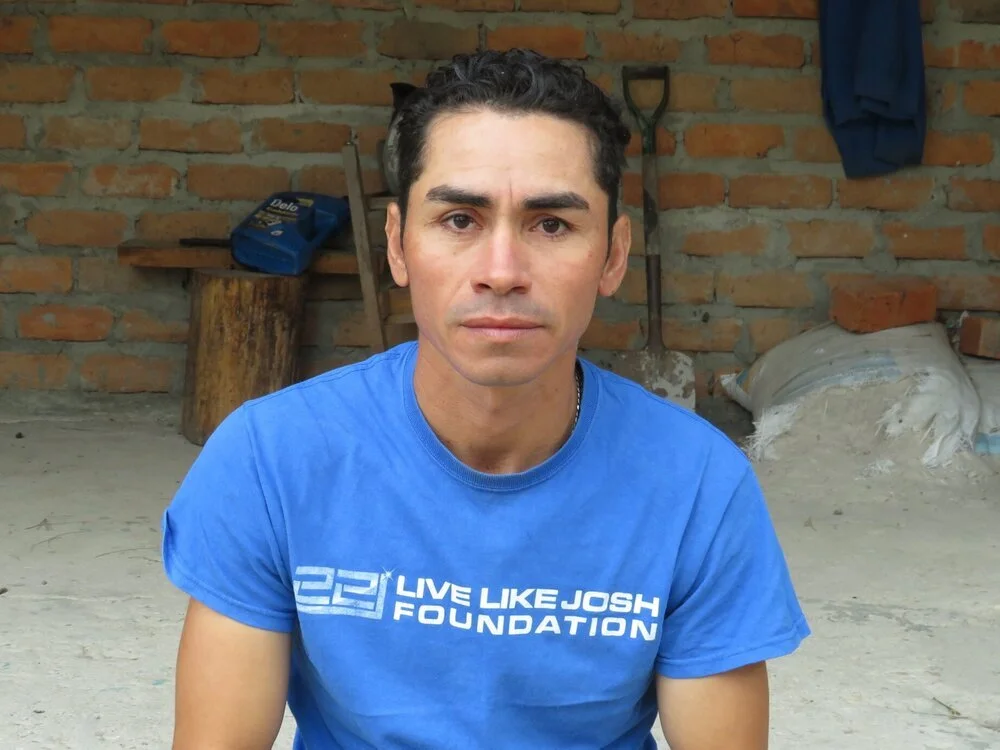 Man wearing a blue 'Live Like Josh Foundation' T-shirt, sitting in front of a brick wall, with tools and construction materials in the background.