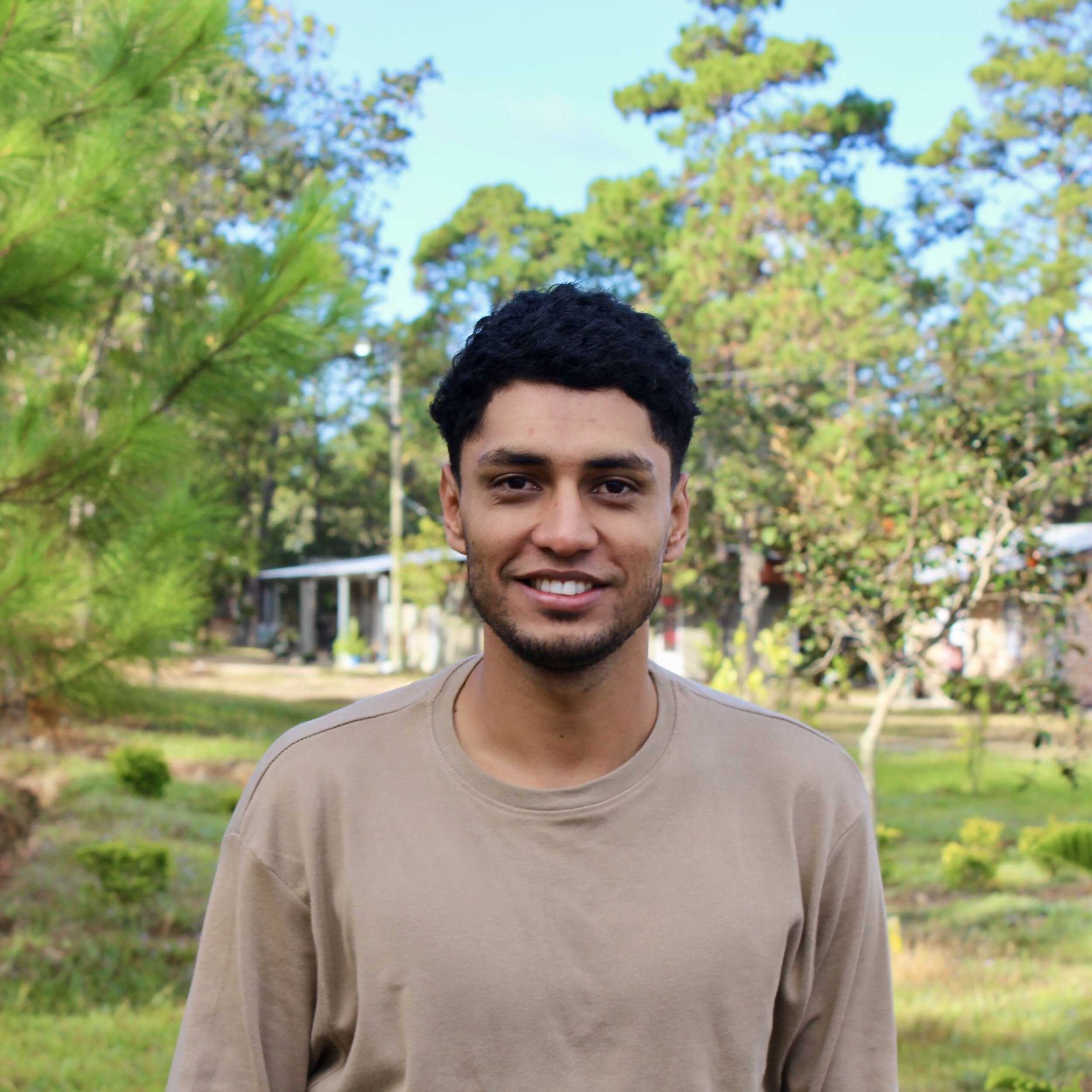 Portrait of a young man with short dark hair smiling outdoors on a sunny day, with trees and a blue sky in the background.