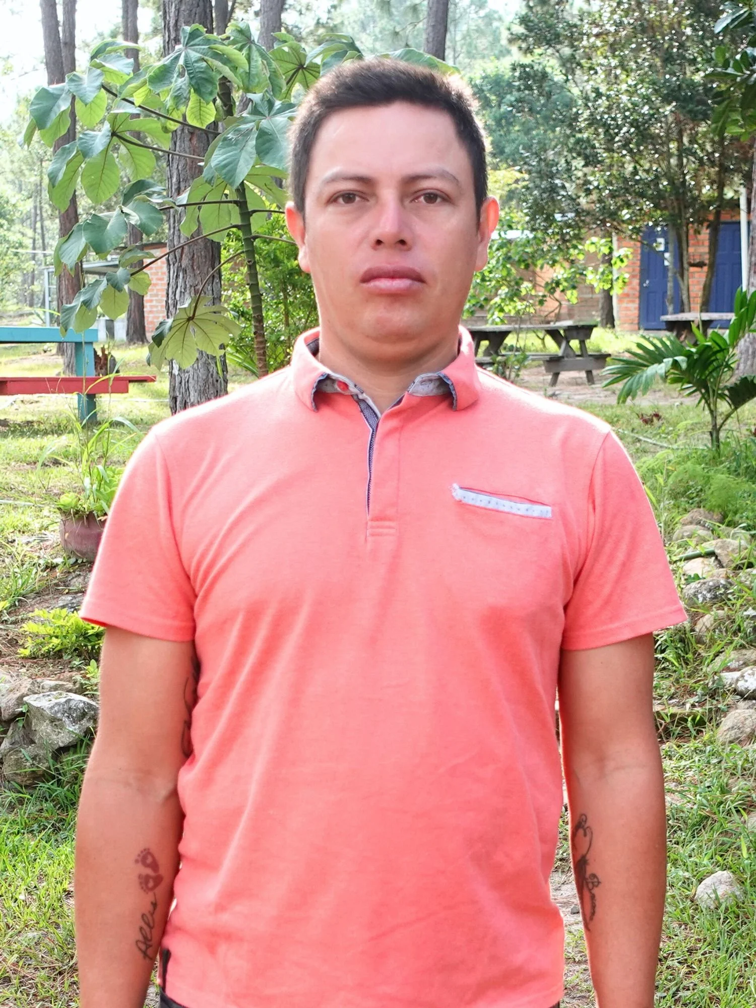 A young man stands outdoors in a lush, green garden with trees, plants, and a picnic table in the background. He has short dark hair, is wearing a salmon-colored shirt, and has tattoos on both forearms.