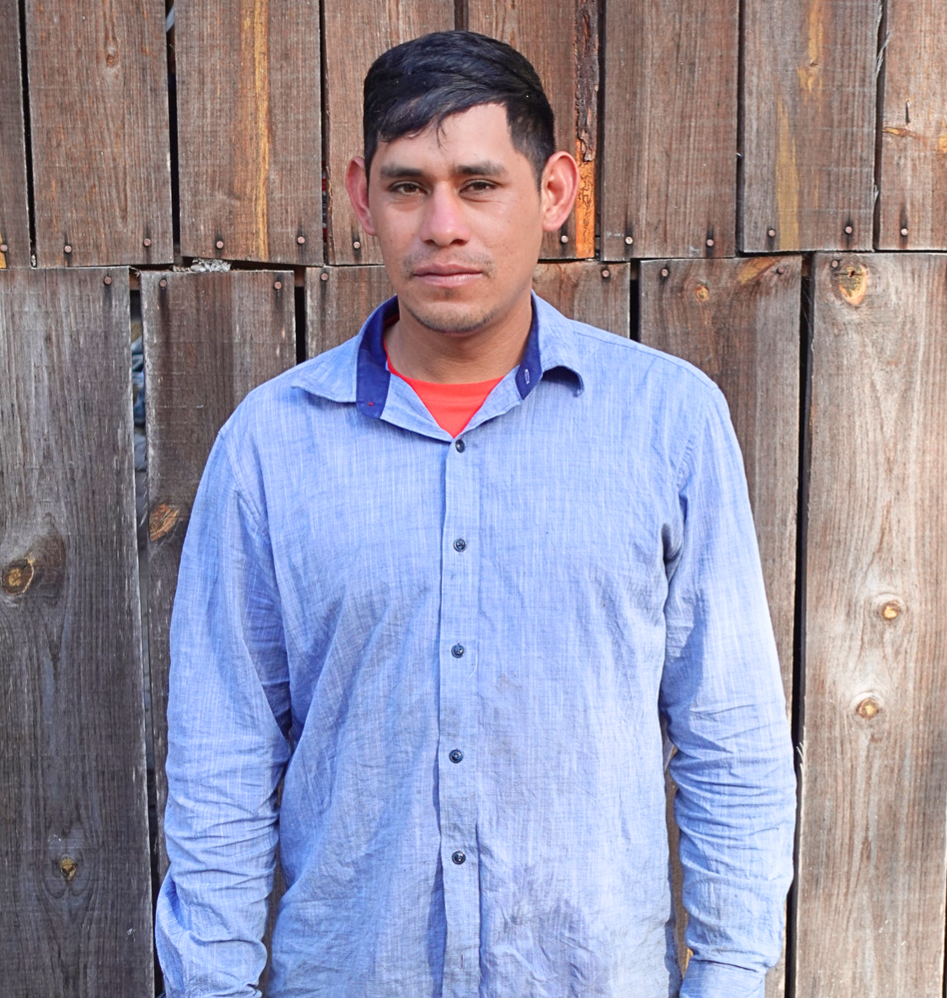 A man with short dark hair wearing a light blue button-up shirt over a red shirt, standing in front of a wooden fence.