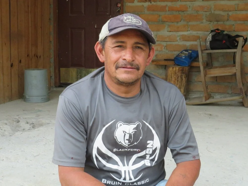 Man wearing a gray t-shirt and a baseball cap, sitting outdoors in front of a brick wall.