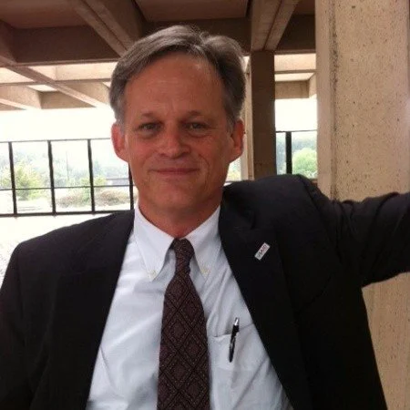A middle-aged man in a suit and tie smiling at camera inside a building with large windows and exposed ceiling beams.