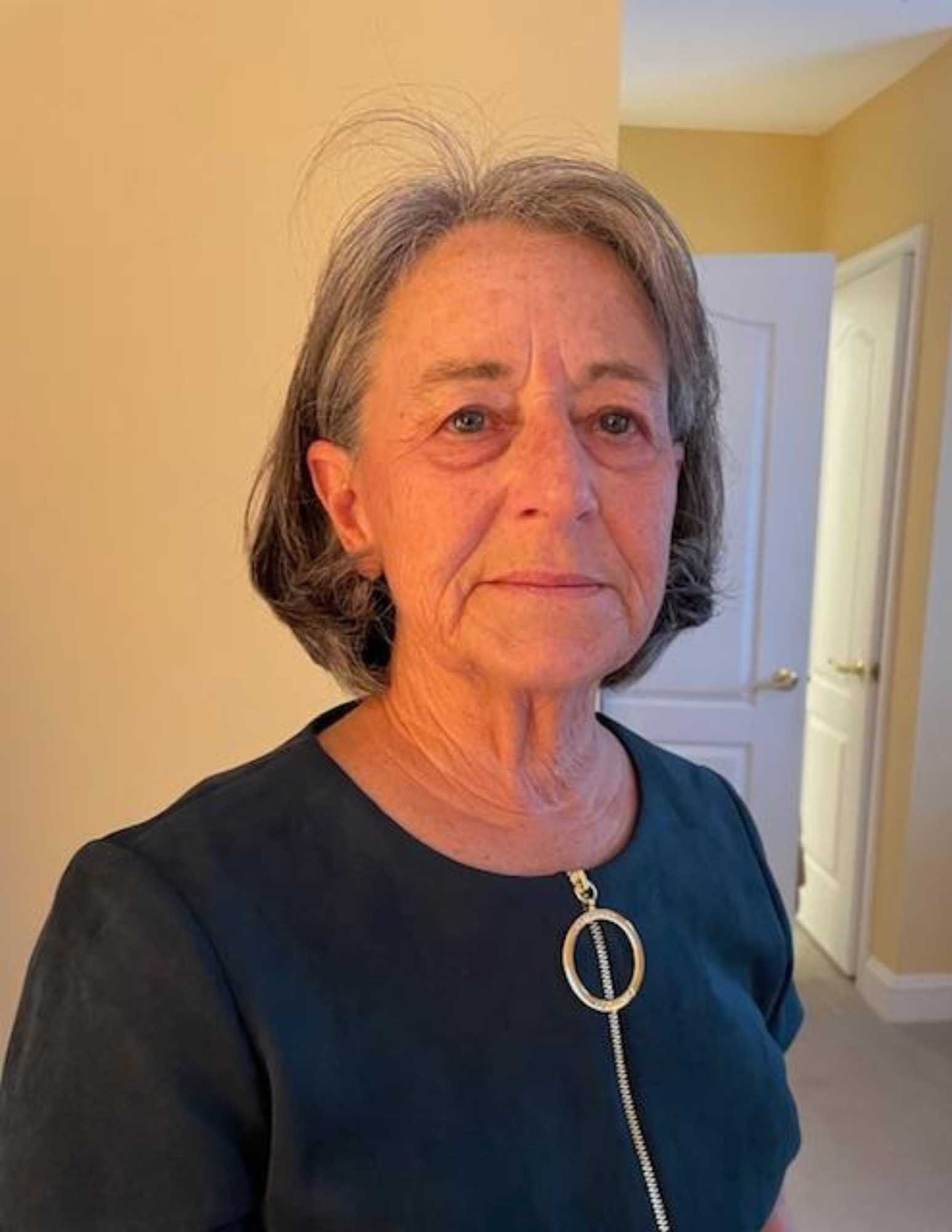Close-up of an older woman with gray hair, wearing a black top with a silver zipper and a large silver ring necklace, standing in a yellow-painted room with an open door in the background.