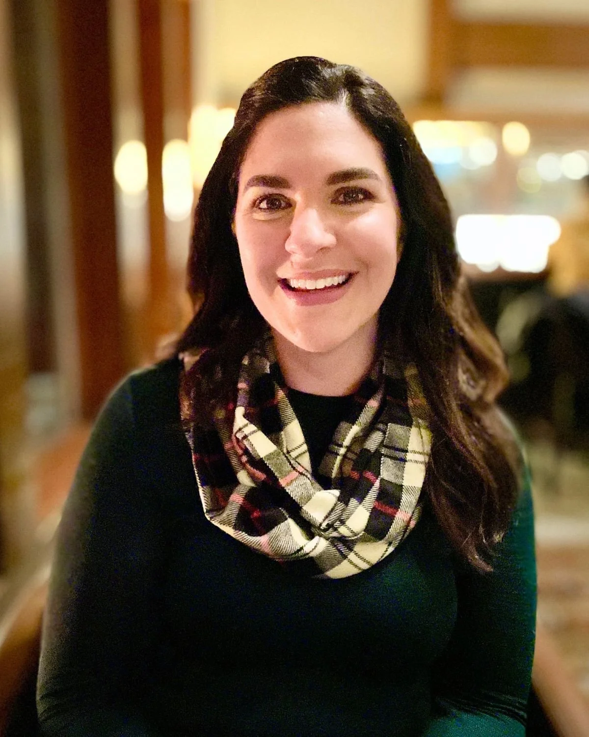 A woman with dark hair, smiling, wearing a black top and a plaid scarf, in a warmly lit indoor setting.